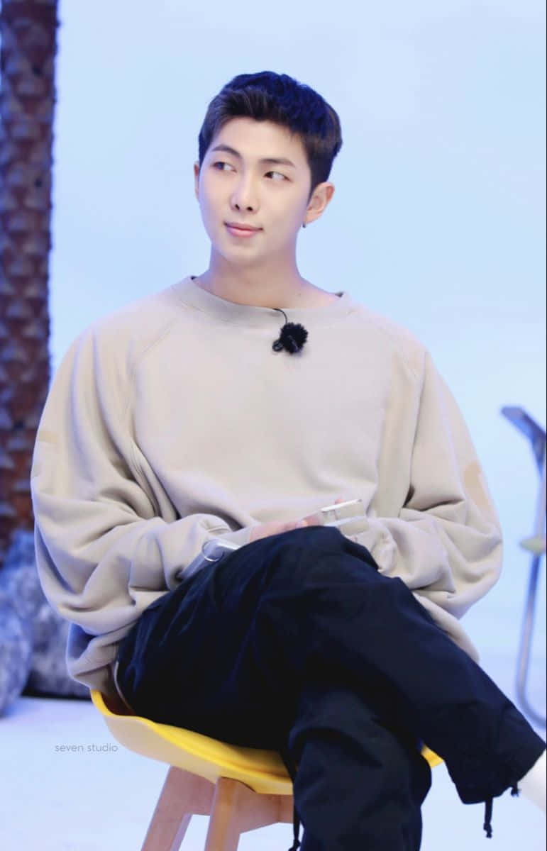 Bts Endorsements 桌布 壁紙 — 一位身穿米色連帽衫的年輕男子，隨意地坐在黃色椅子上，背景是柔和的藍色攝影棚。