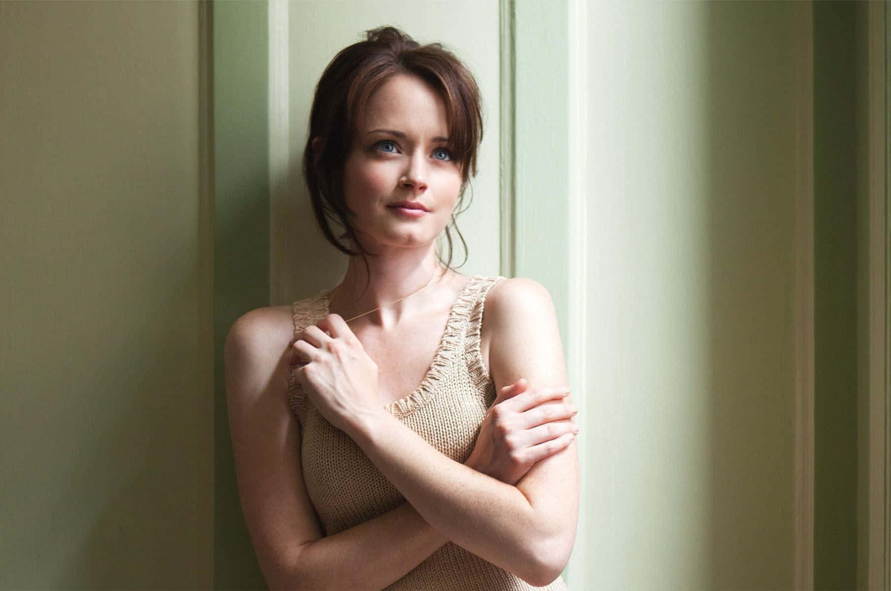 Alexis Bledel 桌布 壁紙 — 一位身穿米色針織上衣的女性站在窗邊，柔和的光線在她臉上和手臂上投下輕柔的陰影。