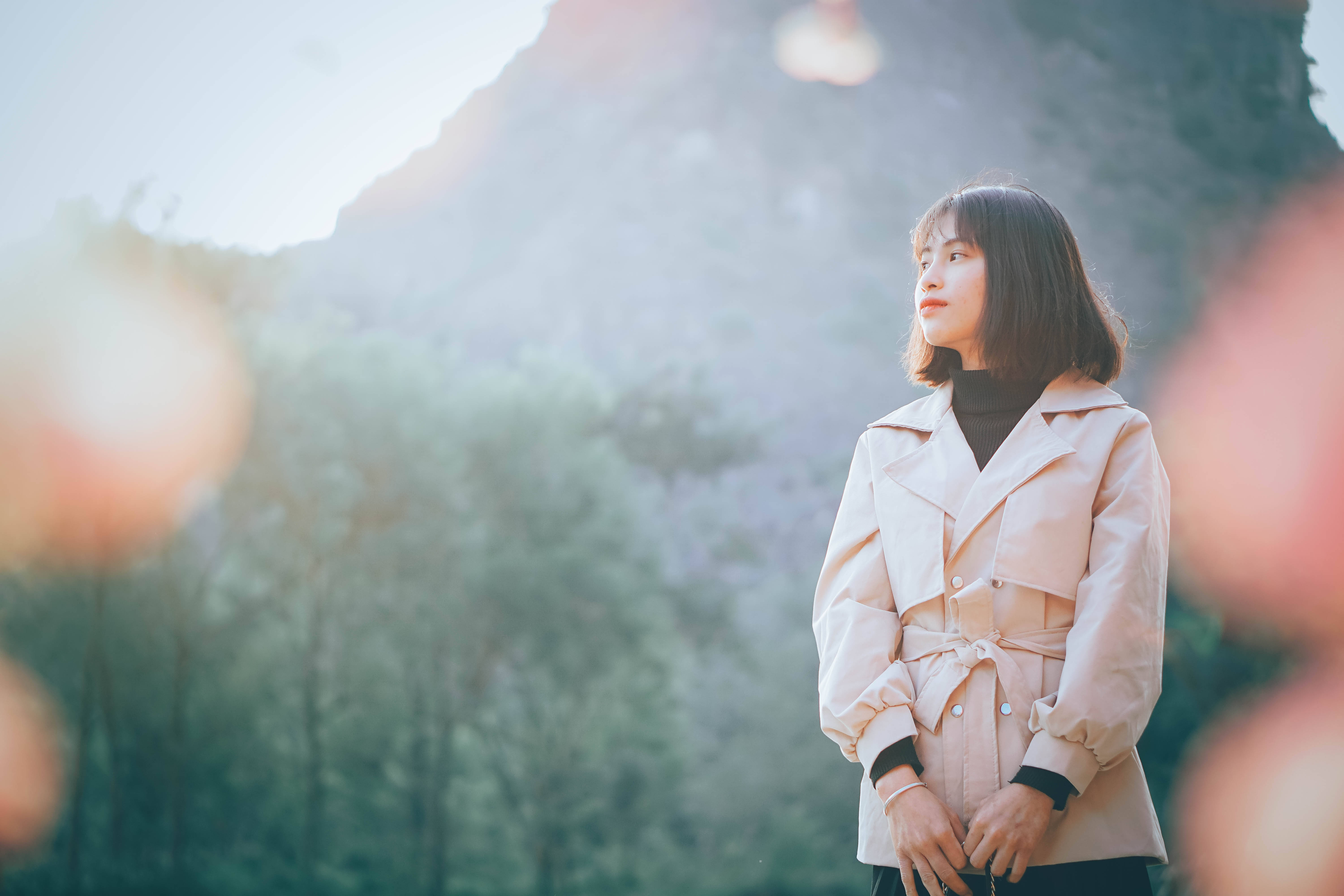 Teenage 桌布 壁紙 — 一位身穿米色風衣的年輕女性，在陽光明媚的森林中沉思，背景是山巒。