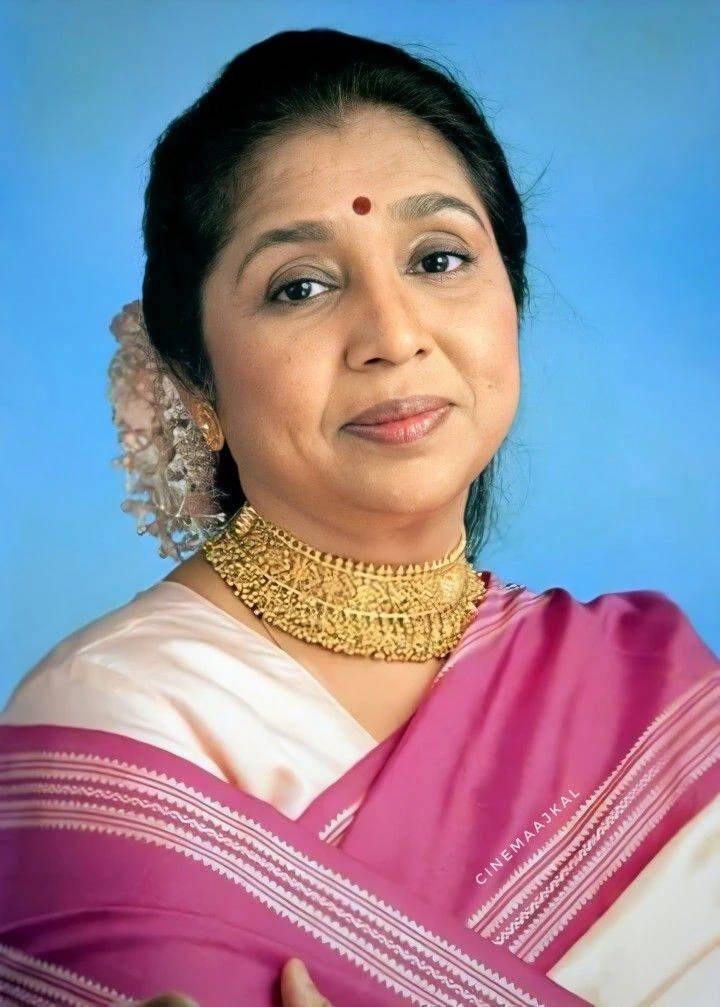 Lata Mangeshkar 桌布 壁紙 — 一位身穿粉白色紗麗、佩戴金飾的優雅女性，在純藍色背景前溫柔微笑。