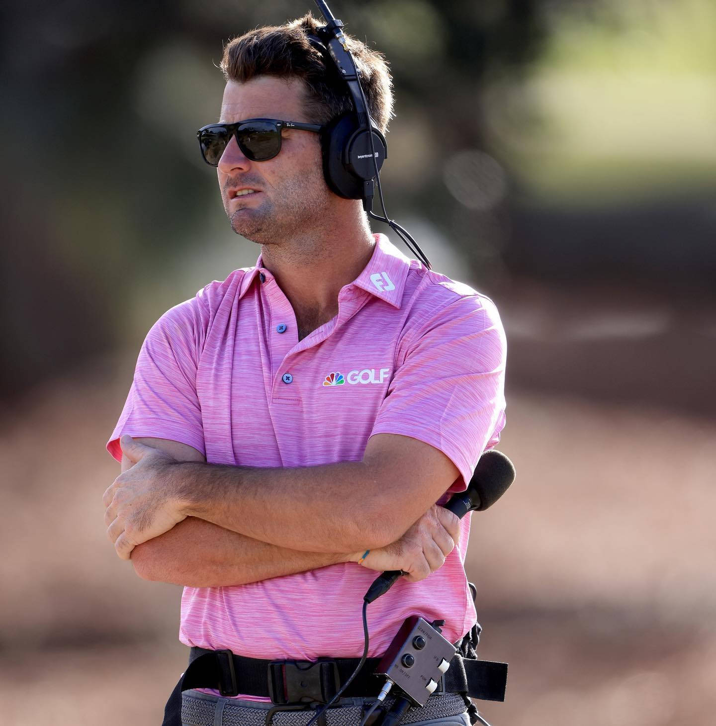 高爾夫 桌布 壁紙 — 一位身穿粉紅色NBC GOLF Polo衫的男性高爾夫球評，雙手交叉，戴著耳機和太陽眼鏡站在戶外。