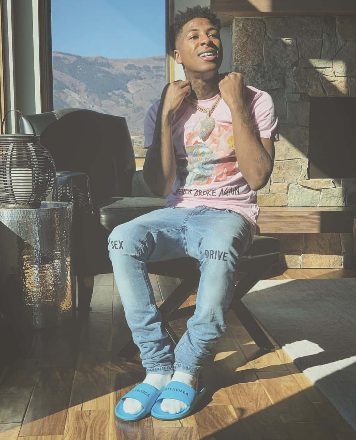 Youngboy 桌布 壁紙 — 一位身穿粉紅色圖案T恤和藍色拖鞋的年輕男子自信地坐在室內的椅子上，微笑著，身後的大窗戶可以看到山景。