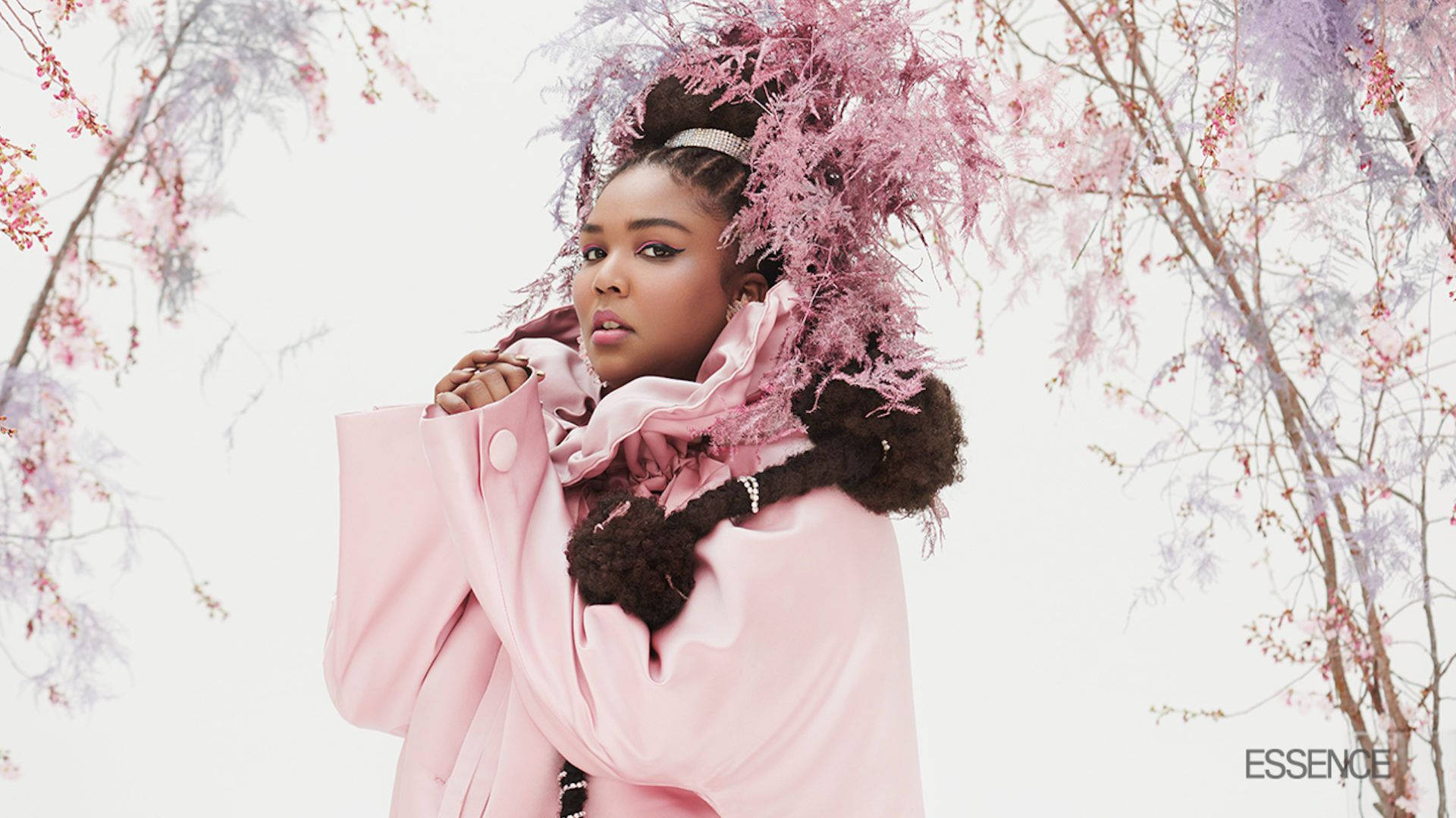 Lizzo 桌布 壁紙 — 一位身穿粉紅色外套和羽毛頭飾的女性，站在盛開的粉紅色和紫色樹木下，背景是柔和的白色天空。