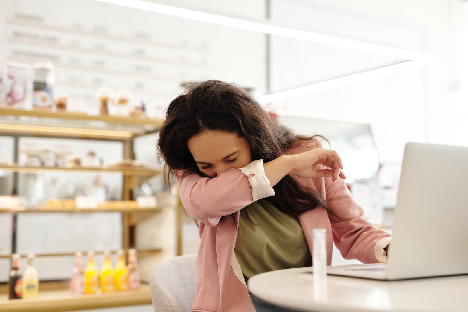 Sneeze 桌布 壁紙 — 一位身穿粉紅色外套的女性在明亮的咖啡館裡，對著筆記型電腦打呵欠，身後架子上擺滿了糕點和飲料。