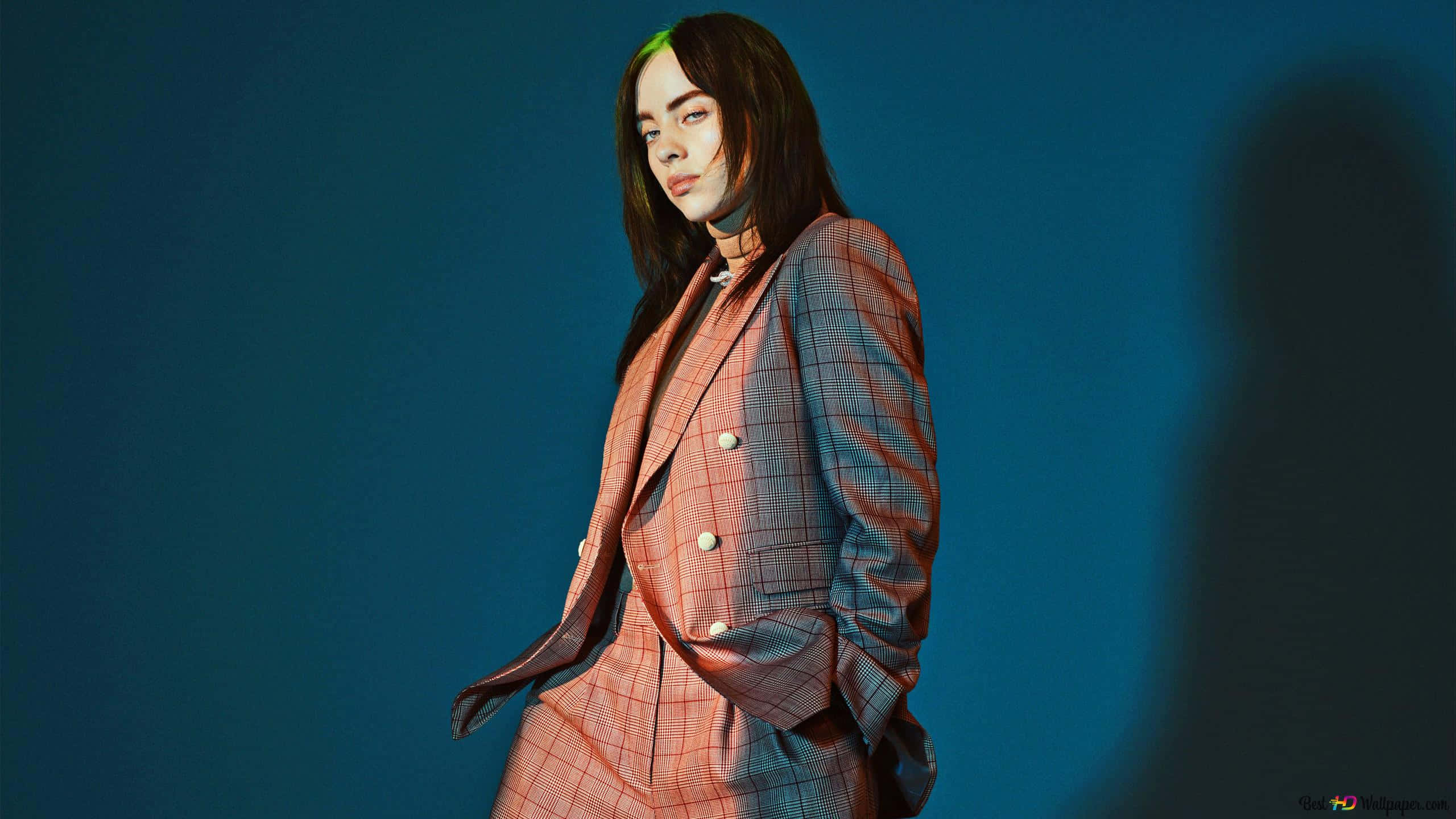 Billie Eilish Laptop 桌布 壁紙 — 一位身穿粉紅色格紋西裝的女性站在深藍色攝影棚背景前，帶有戲劇性的燈光和身後投射的影子。