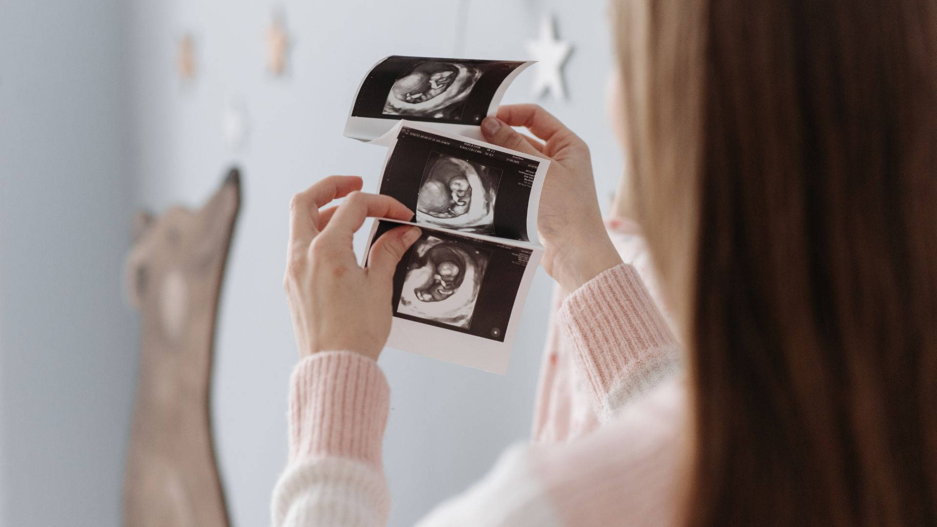 Pregnant 桌布 壁紙 — 一位身穿粉紅色毛衣的女士，手持三張發育中嬰兒的超音波圖像，背景是柔和裝飾的牆壁。