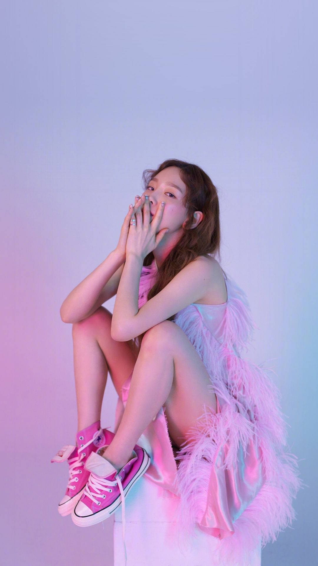 Taeyeon 桌布 壁紙 — 一位身穿粉紅色羽毛服裝和運動鞋的年輕女性，雙腿交叉坐在柔和的漸變背景前，雙手捂住嘴巴，表現出俏皮的驚訝。