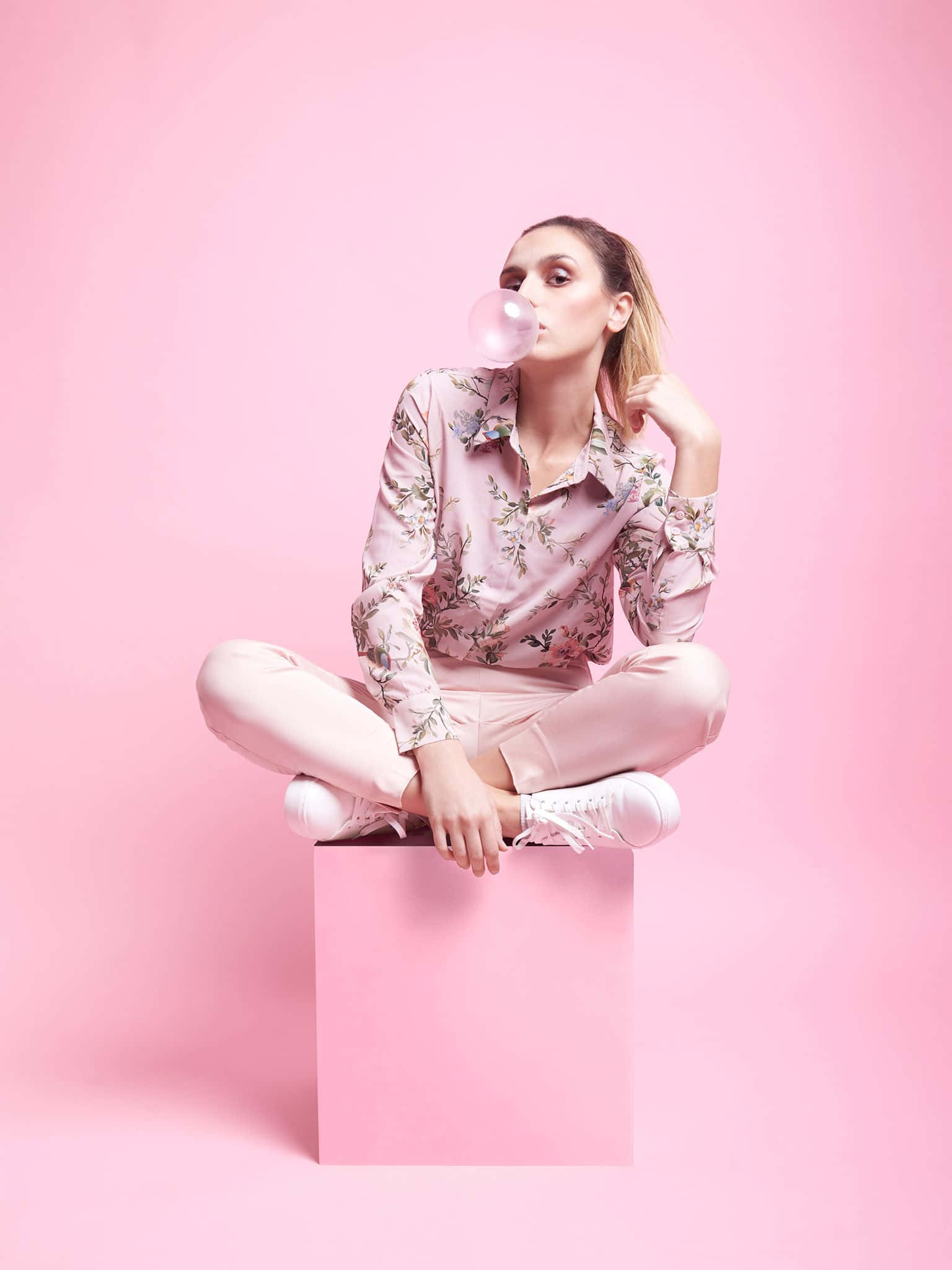 Pink Fashion 桌布 壁紙 — 一位身穿粉紅色花卉服飾的女性，盤腿坐在粉紅色立方體上，對著純粉紅色的攝影棚背景吹泡泡。