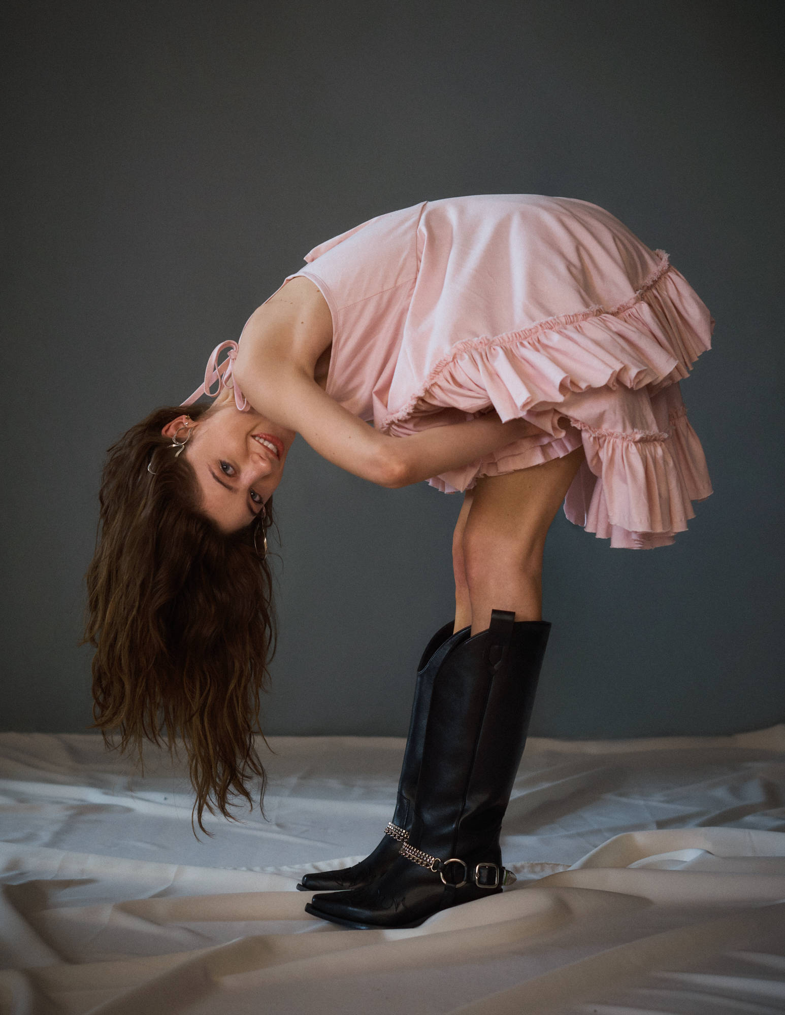 Natalia 桌布 壁紙 — 一位身穿粉紅色荷葉邊連衣裙和黑色靴子的女士，在灰色牆壁前的白色床單上向後彎腰。