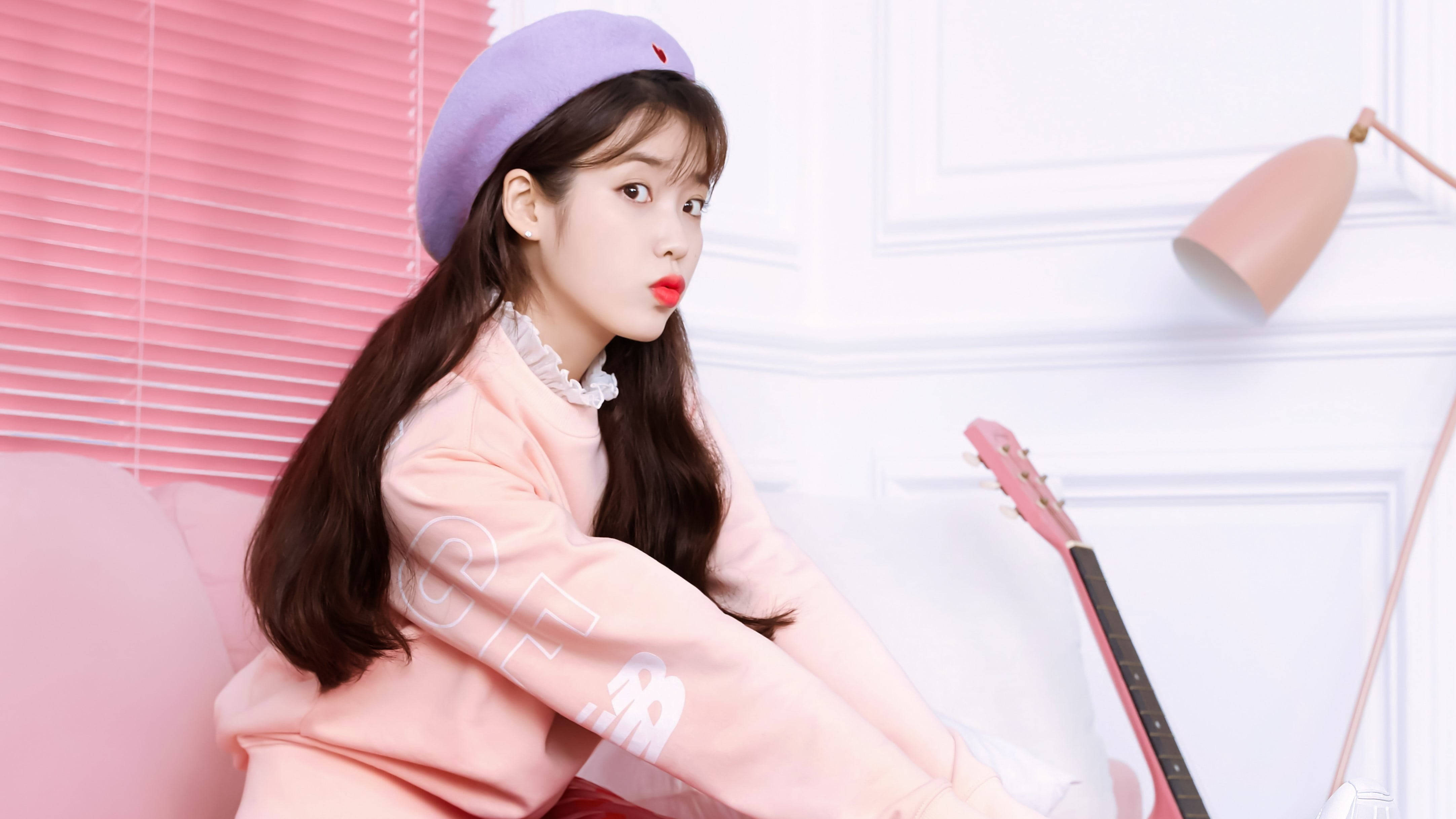 Kpop Aesthetic 桌布 壁紙 — 一位身穿粉紅色衛衣和紫色貝雷帽的年輕女性，在粉彩粉紅和白色的房間裡，拿著吉他擺出俏皮的姿勢。