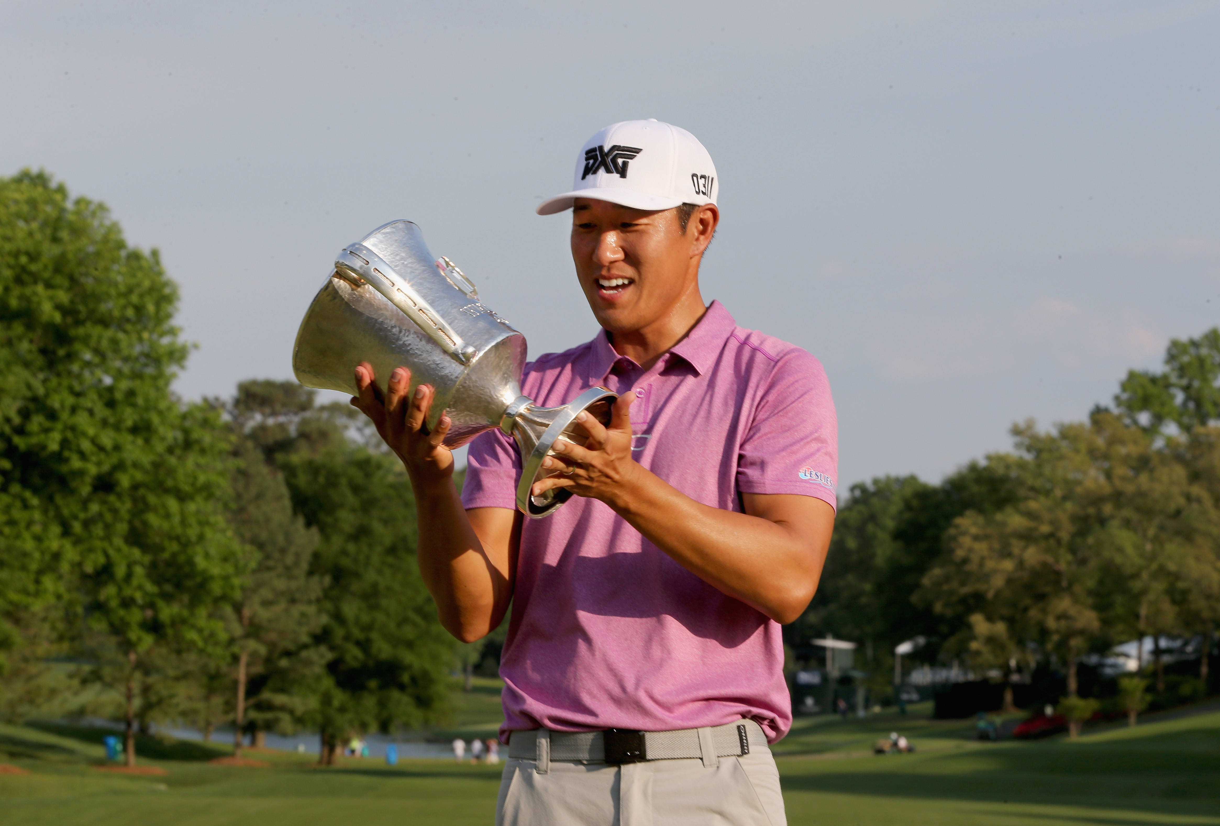 Trophy 桌布 壁紙 — 一位身穿粉紅色襯衫、面帶微笑的高爾夫球手，在陽光明媚、綠樹成蔭的高爾夫球場上自豪地高舉一個大型銀色獎盃。