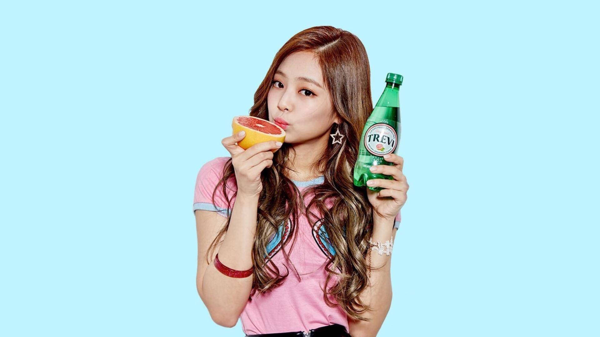 Jennie 桌布 壁紙 — 一位身穿粉紅色襯衫的年輕女性，手持綠色Trevi汽水瓶，正在吸吮葡萄柚，背景為純淺藍色。