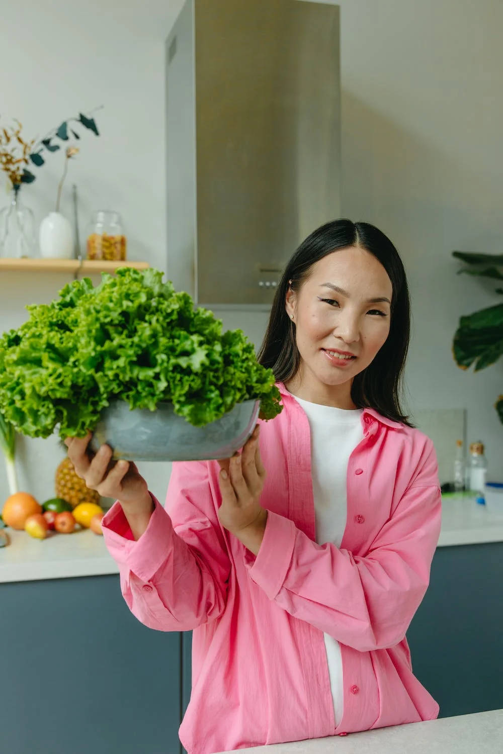 Vegetables 桌布 壁紙 — 一位身穿粉紅色襯衫的微笑女士，在現代廚房裡自豪地捧著一大碗新鮮的綠色生菜。