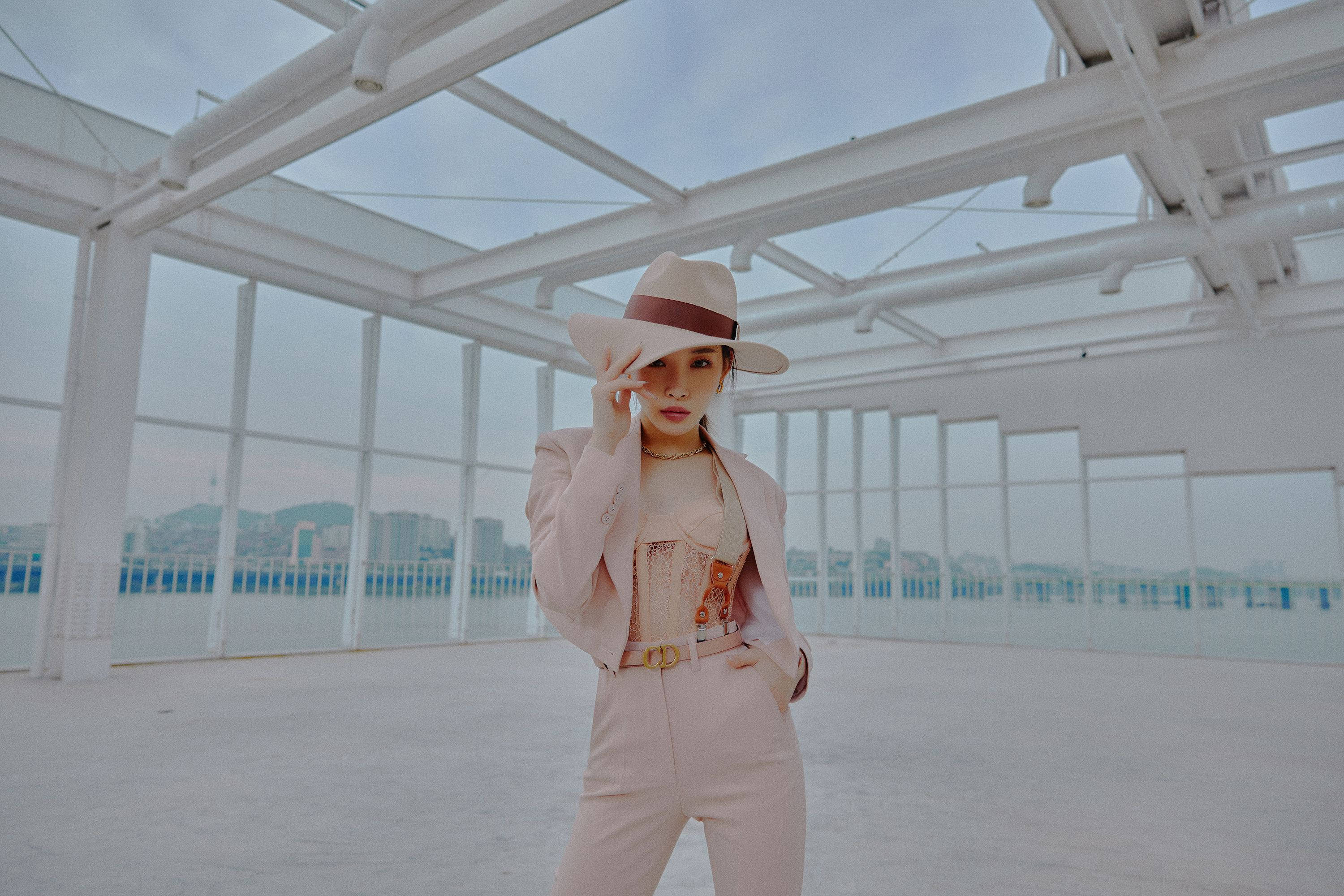 Chungha 桌布 壁紙 — 一位身穿粉紅色西裝和帽子的女性，在一個現代化的玻璃屋頂天台上自信地擺姿勢，俯瞰城市景觀。