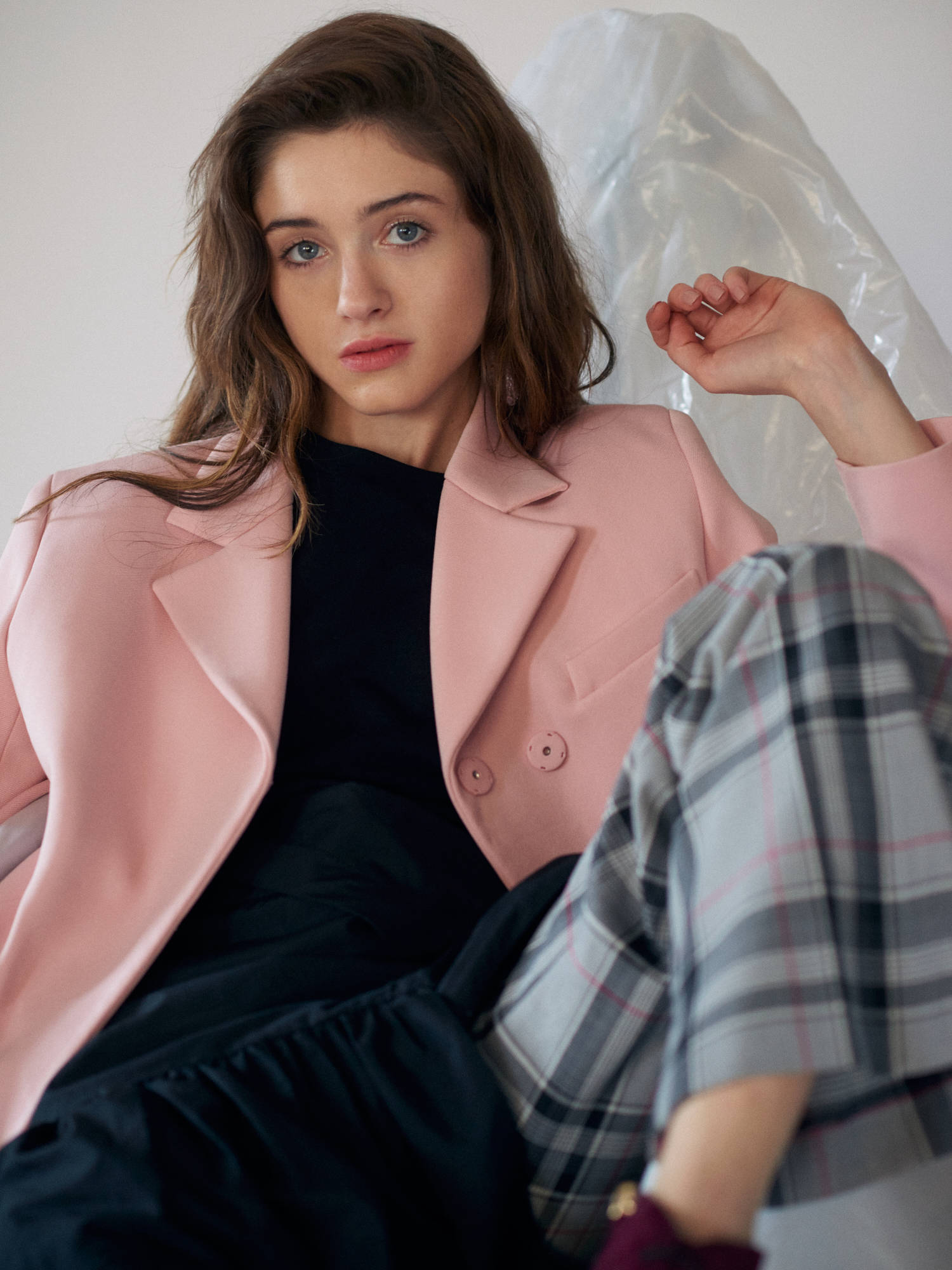 Natalia 桌布 壁紙 — 一位身穿粉紅色西裝外套和格子裙的年輕女性，靠著白色牆壁坐著，身後有一個塑膠袋。