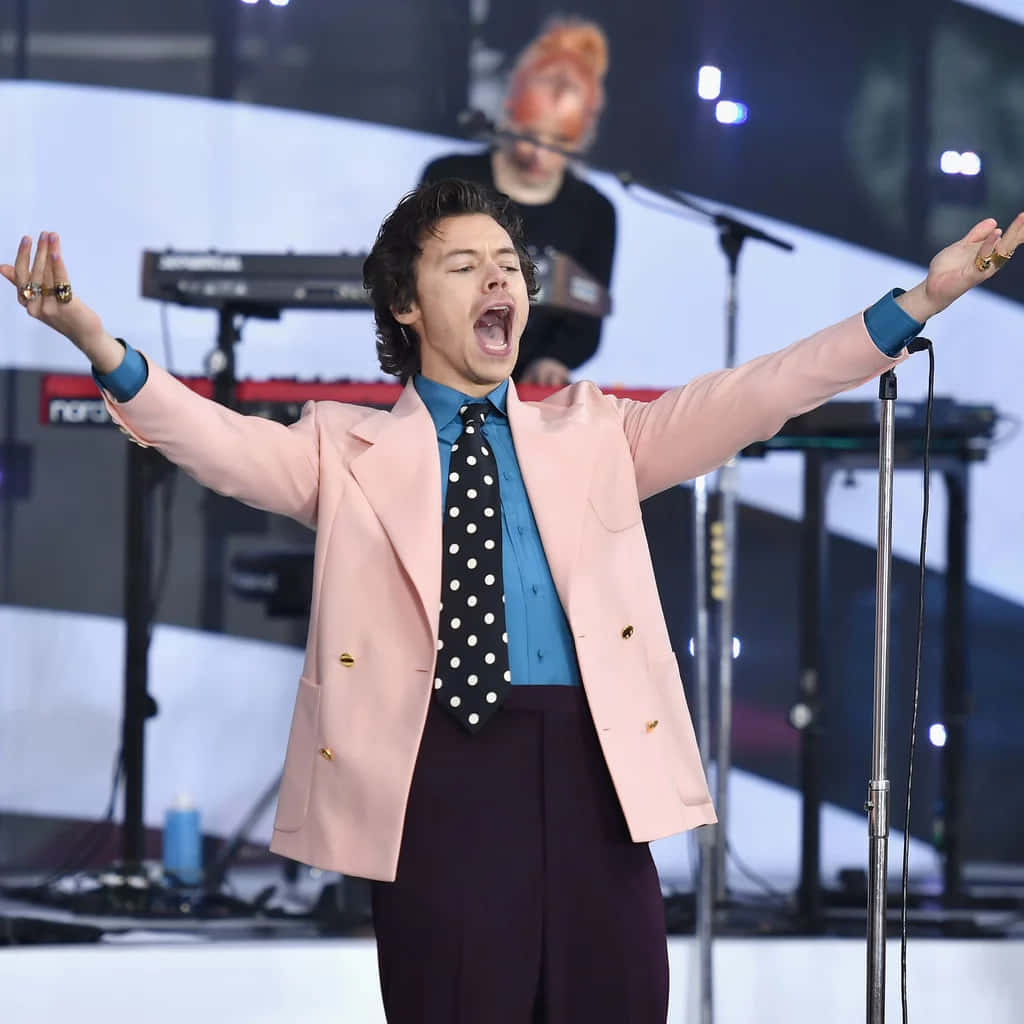 Harry Styles 2021 桌布 壁紙 — 一位身穿粉紅色西裝外套和波點領帶的男歌手，在燈光明亮的舞台上張開雙臂熱情表演。