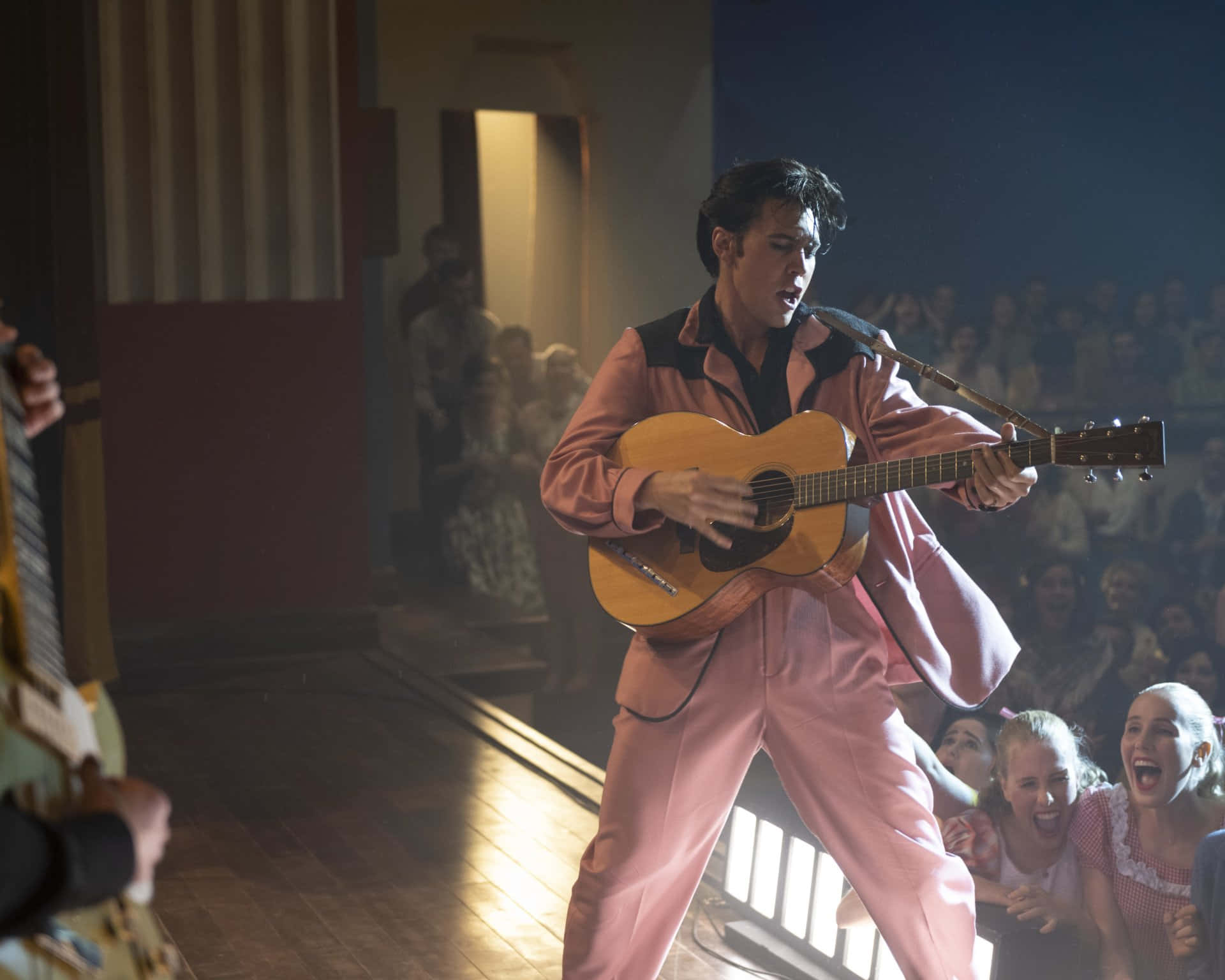 4K Elvis 桌布 壁紙 — 一位身穿粉紅色西裝的年輕男子在溫暖的聚光燈下熱情地彈奏吉他，興奮的群眾歡呼喝采。