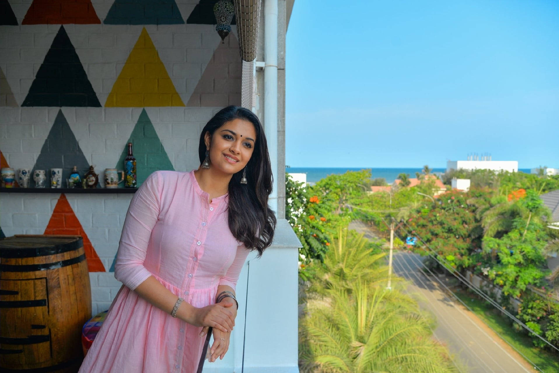 telugu 桌布 壁紙 — 一位身穿粉紅色連衣裙的女性在陽台上微笑，俯瞰著晴朗藍天下鬱鬱蔥蔥的熱帶海岸景色。