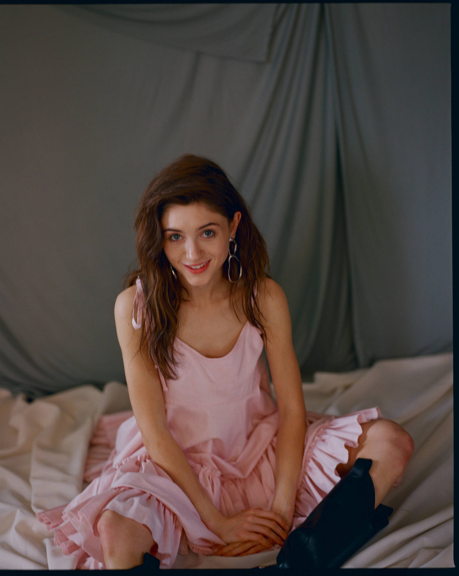 Natalia 桌布 壁紙 — 一位身穿粉紅色連衣裙的年輕女性坐在床上，身後有灰色布料垂墜，露出溫暖的笑容。