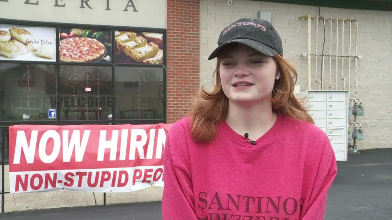 Stupid 桌布 壁紙 — 一位身穿粉紅色運動衫和帽子的年輕女子站在一家披薩店前微笑，店內掛著「NOW HIRING NON-STUPID PEOPLE」的牌子。