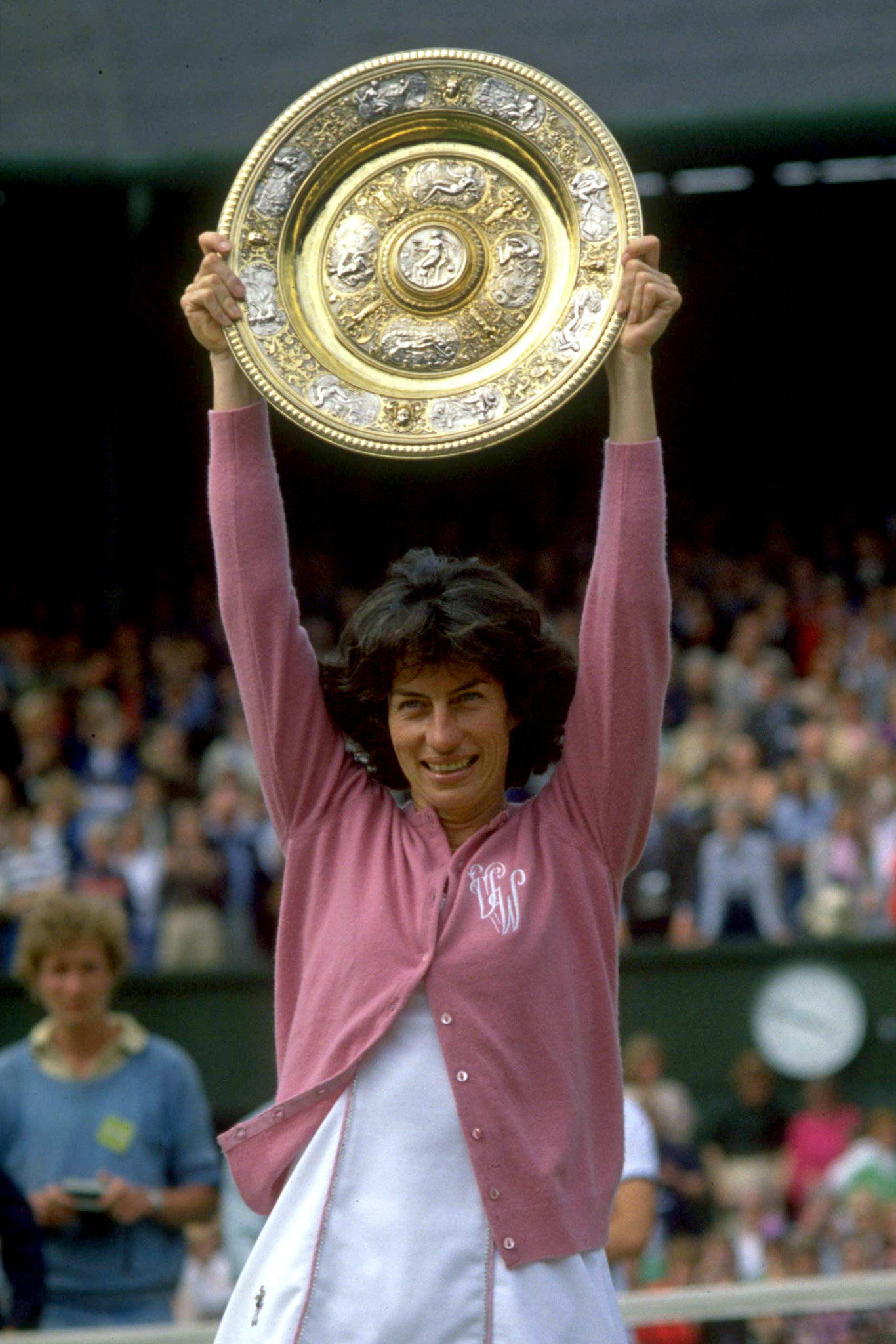 Tennis 桌布 壁紙 — 一位身穿粉紅色開襟毛衣的快樂網球選手，在草地上高舉一個華麗的大型金色獎盃，慶祝勝利。