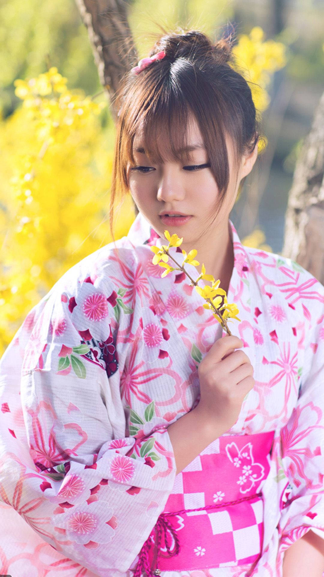 Cute Girl 桌布 壁紙 — 一位身穿粉紅花卉和服的年輕女子，在陽光明媚的黃色花叢前手持一枝黃色花枝。