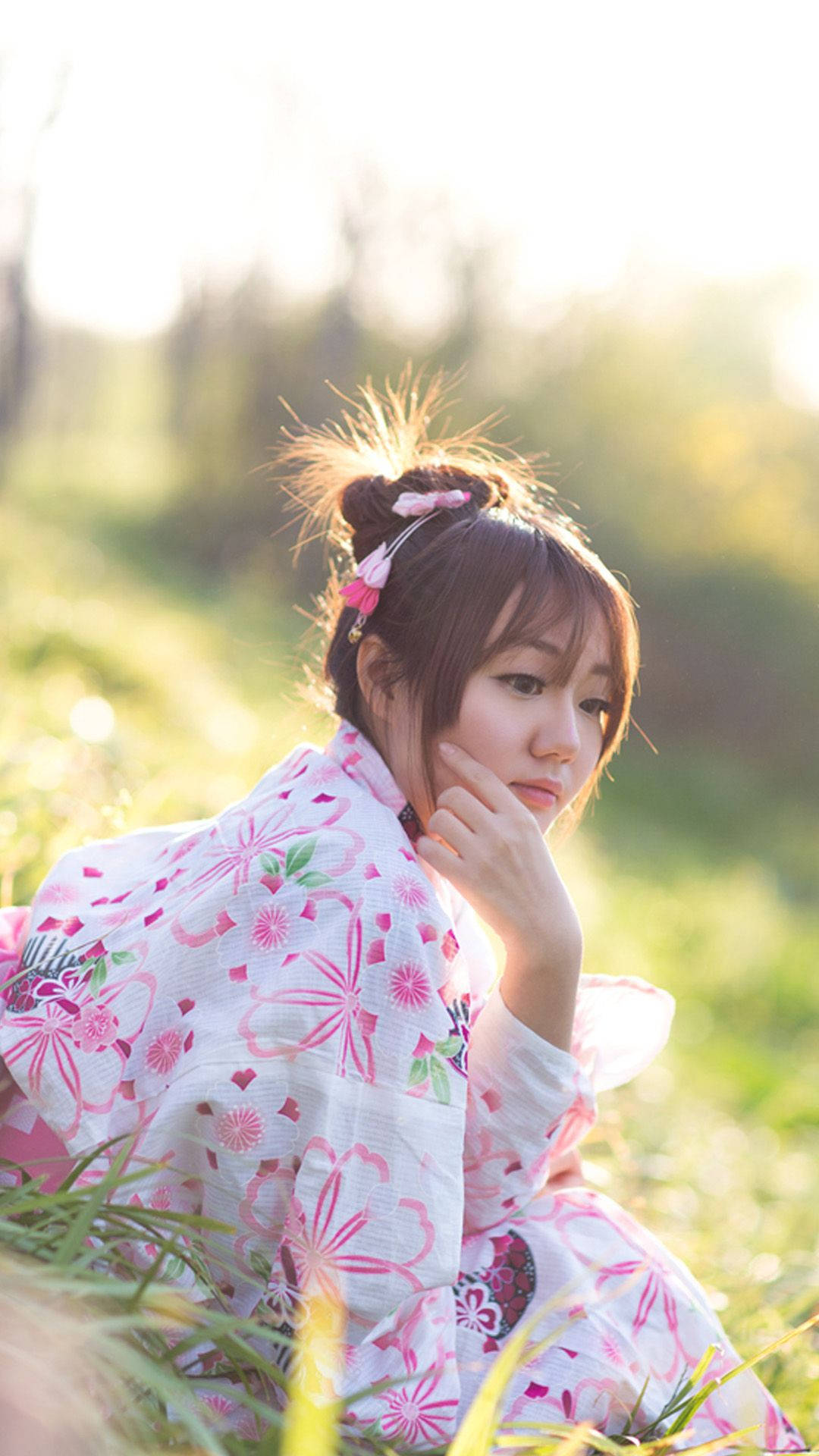 美女 桌布 壁紙 — 一位身穿粉紅花卉和服的年輕女子坐在陽光普照的田野中，她的手輕輕觸碰臉頰，金色的光線從她身後的草叢中透出。