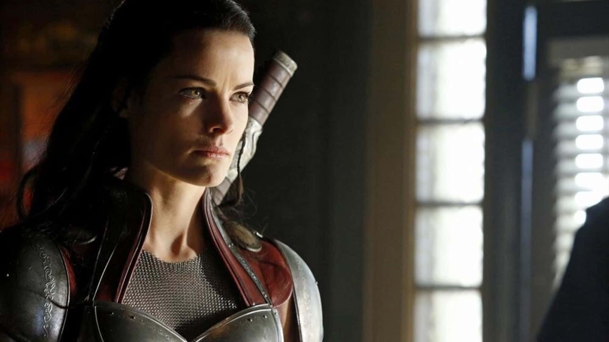 Lady Sif 桌布 壁紙 — 一位身穿精細盔甲的女戰士站在光線昏暗的房間中，陽光從她身後的窗戶照射進來。