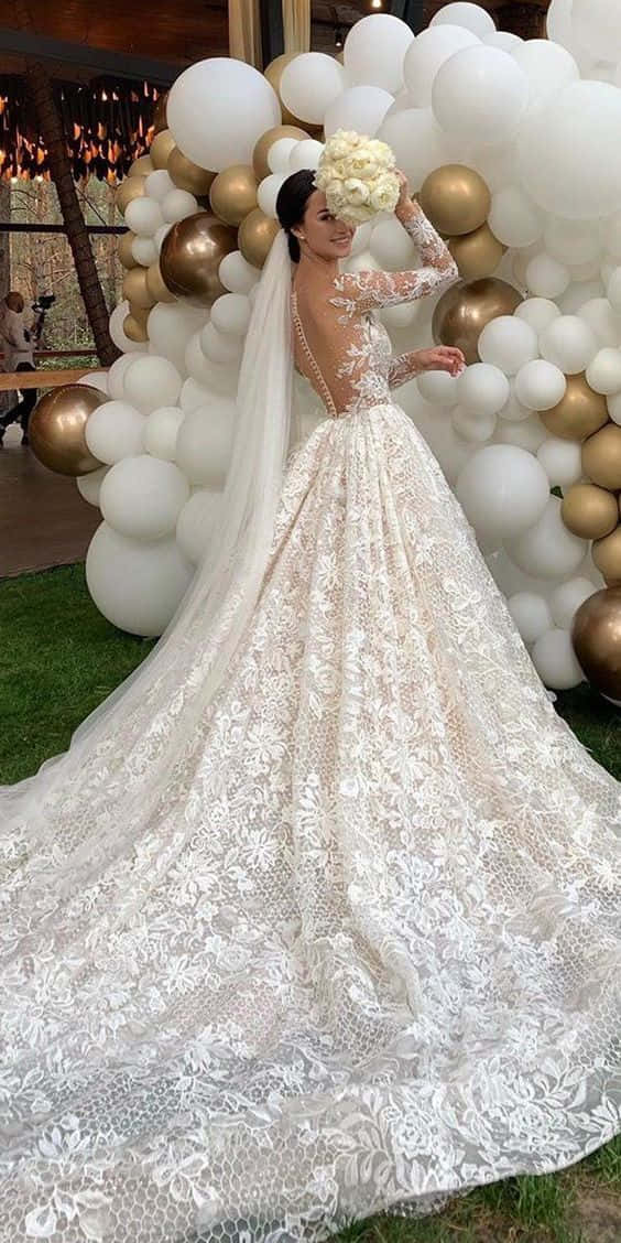 Wedding Gown 桌布 壁紙 — 一位身穿精緻白色蕾絲婚紗、長長拖尾的新娘，站在白色和金色氣球牆前，手持花束。