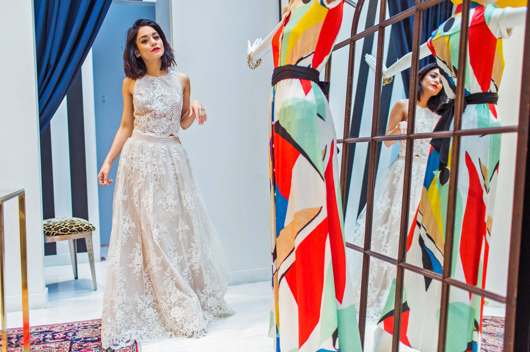 Vanessa Hudgens 桌布 壁紙 — 一位身穿精緻白色蕾絲長裙的女性站在人體模型旁，人體模型上穿著一件充滿活力的抽象連衣裙，映照在時尚精品店的網格框架鏡子中。