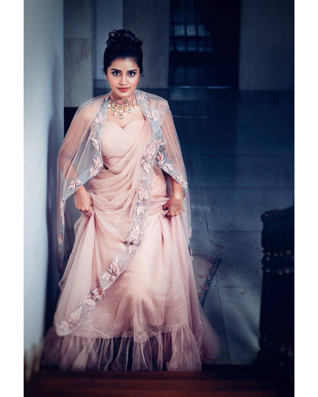 anupama parameswaran 桌布 壁紙 — 一位身穿精緻粉紅色刺繡長裙（Lehenga）搭配透明披肩（Dupatta）的女性，在燈光昏暗的走廊中優雅站立。