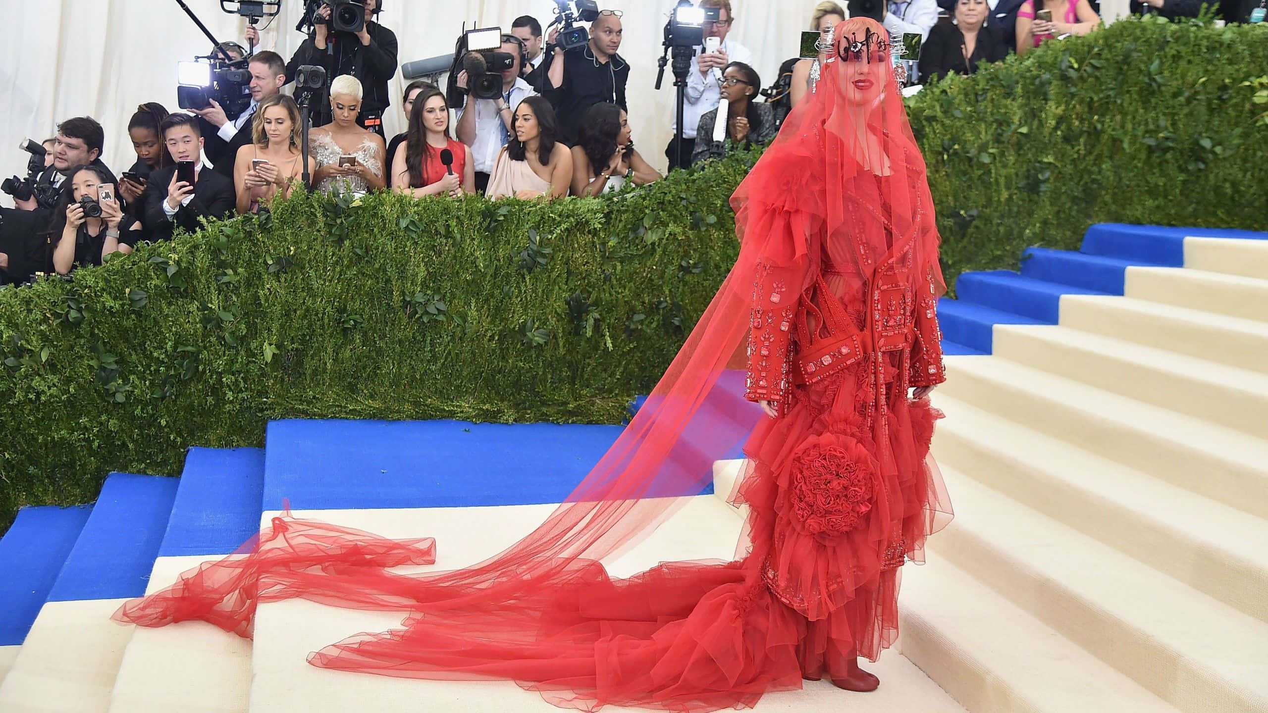 Met Gala 桌布 壁紙 — 一位身穿精緻紅色長裙、披著飄逸頭紗的女性，在一個被攝影師包圍的華麗活動中，走下鋪著藍色地毯的樓梯。