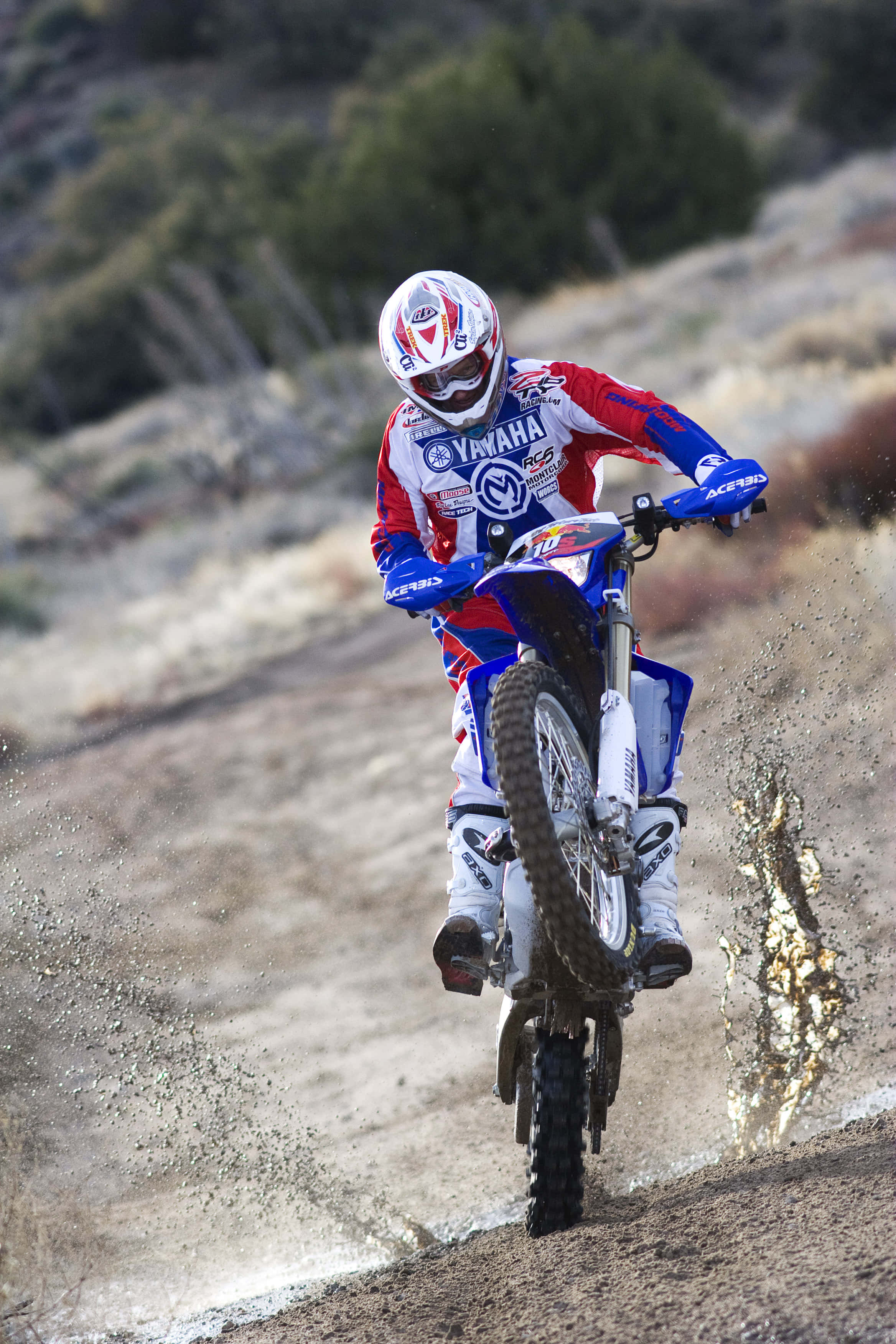 Dirtbike 桌布 壁紙 — 一位身穿紅、藍、白色裝備的越野摩托車手在崎嶇小徑上傾斜轉彎，揚起塵土。