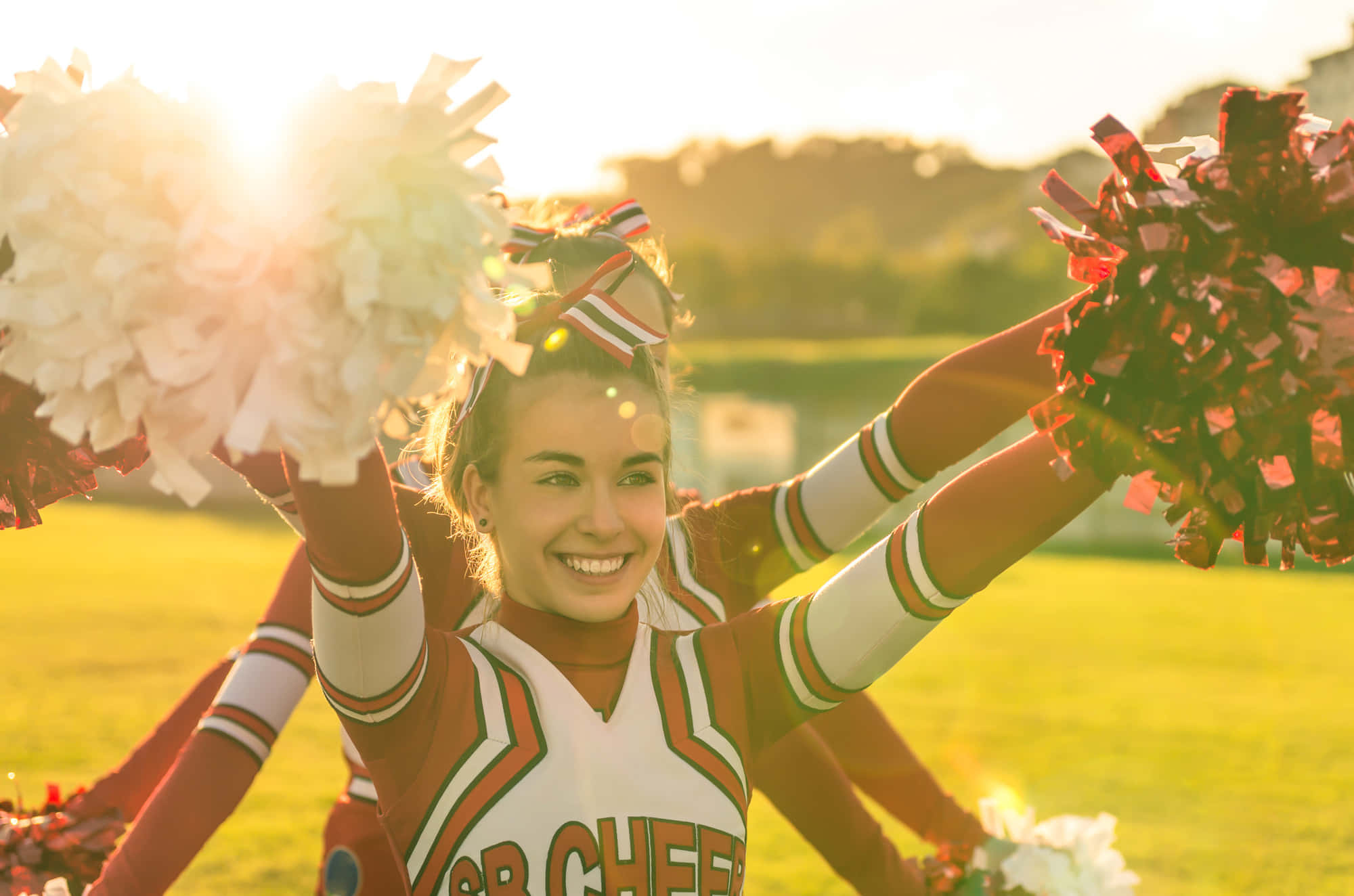 Cheer 桌布 壁紙 — 一位身穿紅白制服、面帶微笑的啦啦隊員，在金色夕陽下的草地上高舉彩球。