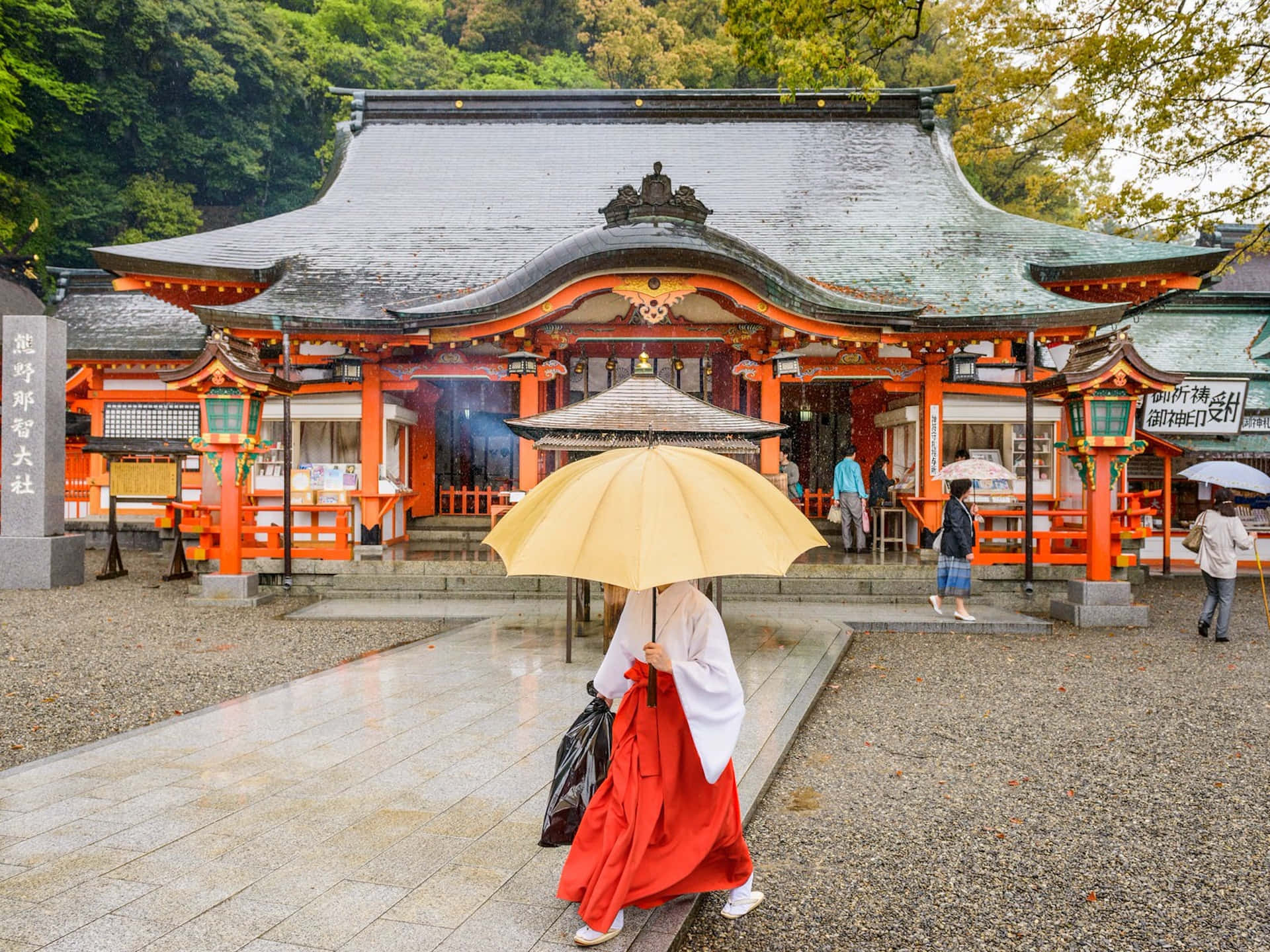 日式 桌布 壁紙 — 一位身穿紅白和服的人撐著黃色雨傘，走向一座擁有橙色柱子和綠色屋頂的傳統日本神社。