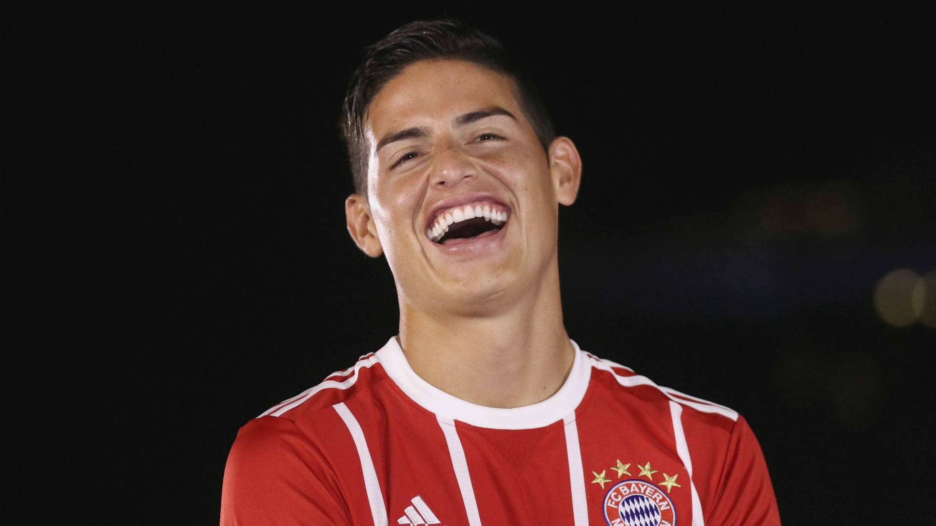 James Rodriguez 桌布 壁紙 — 一位身穿紅白條紋球衣的快樂足球運動員，在黑暗的夜晚背景下，伴隨著散景燈光，開懷大笑。
