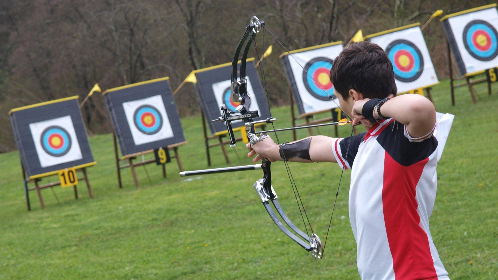 Archery 桌布 壁紙 — 一位身穿紅白黑襯衫的年輕弓箭手拉開現代複合弓，瞄準草地上的目標。