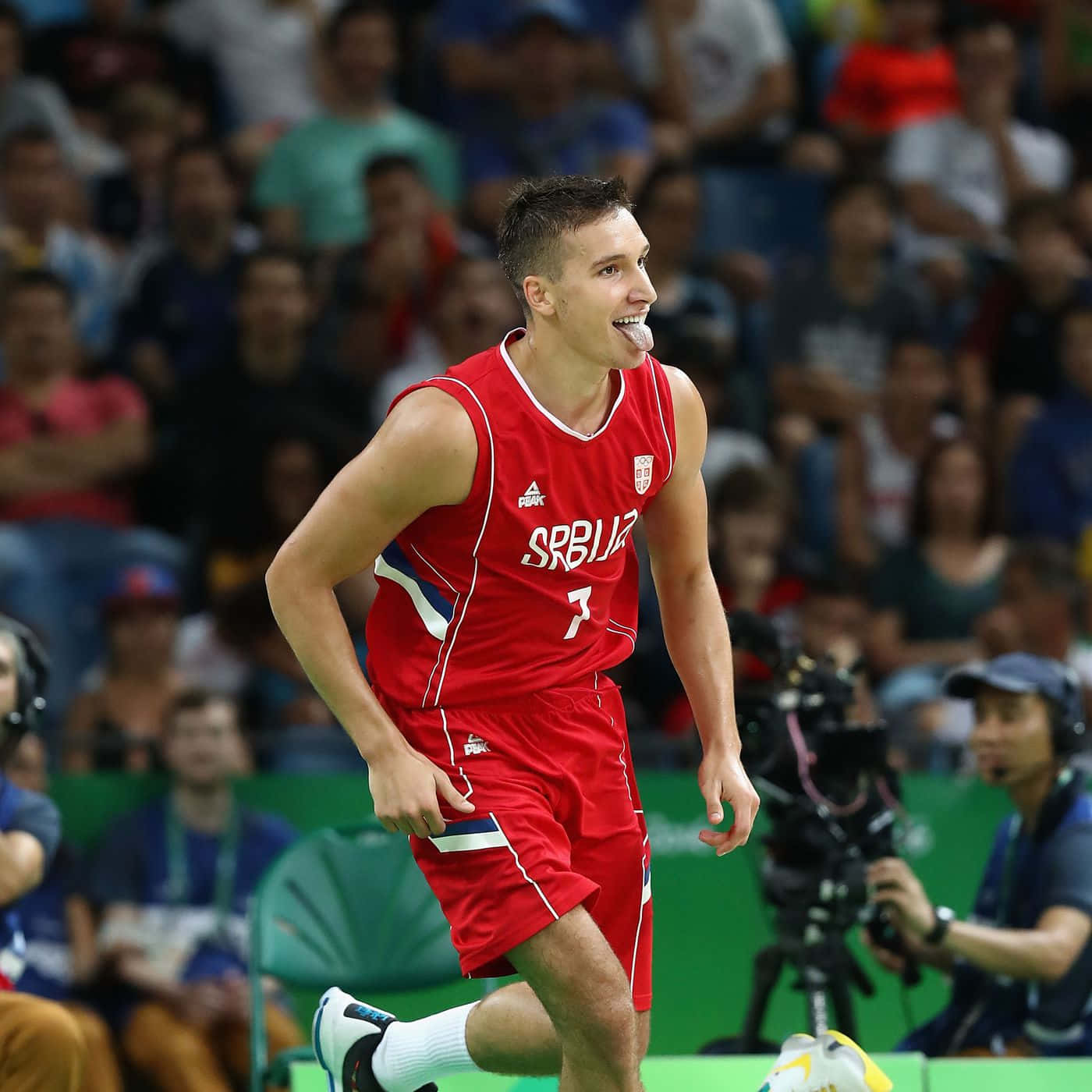 NBA 桌布 壁紙 — 一位身穿紅色7號球衣的塞爾維亞籃球運動員在比賽中頑皮地伸出舌頭，背景是模糊的觀眾。