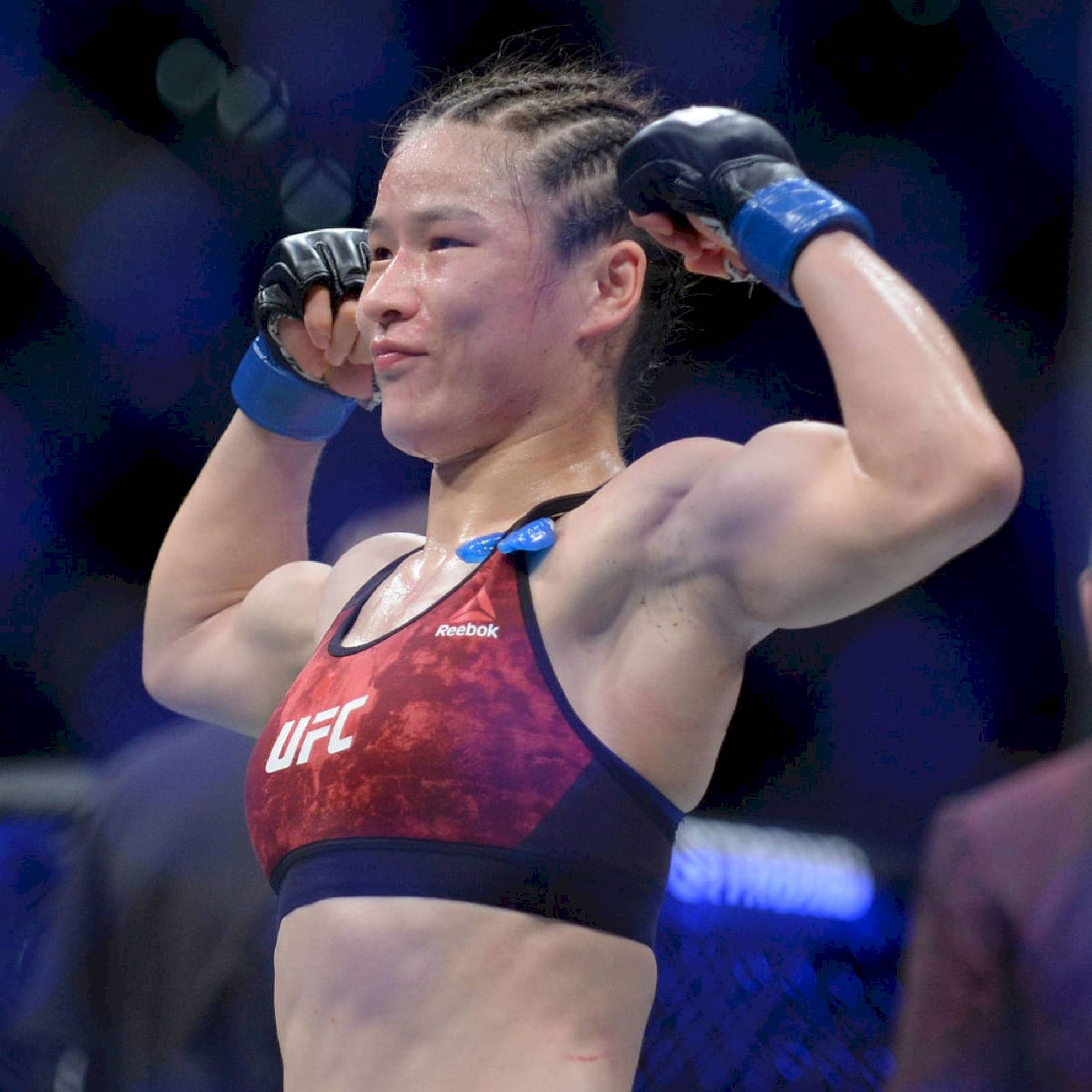Athletic 桌布 壁紙 — 一位身穿紅色Reebok運動胸罩的勝利女UFC拳手，在明亮的場館燈光下於八角籠內高舉戴著手套的拳頭慶祝勝利。