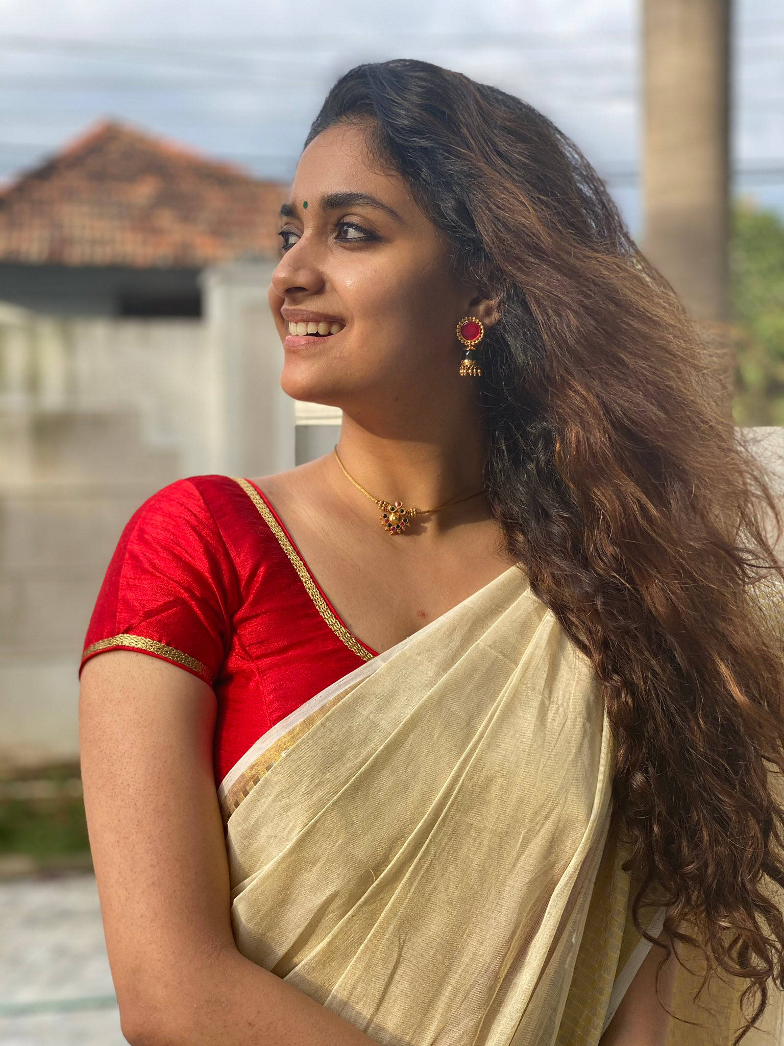 Indian 桌布 壁紙 — 一位身穿紅色上衣和奶油色紗麗的光彩照人的女性，在戶外溫柔微笑，仰望天空。