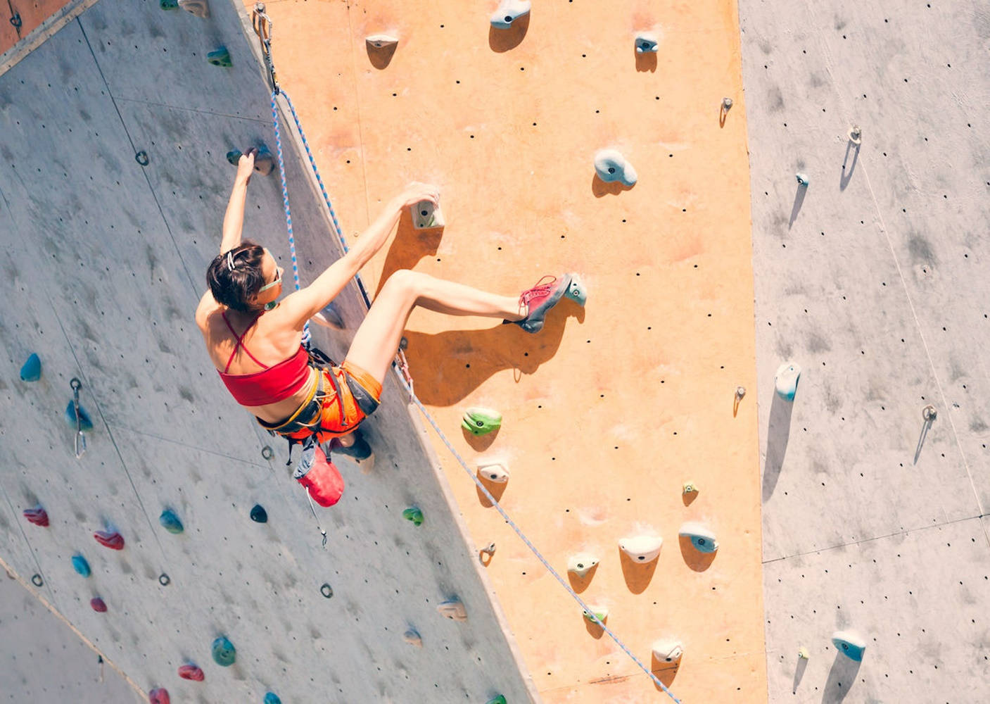 Climbing 桌布 壁紙 — 一位身穿紅色上衣和橙色短褲的女性在明亮的陽光下攀爬色彩繽紛的室內攀岩牆。