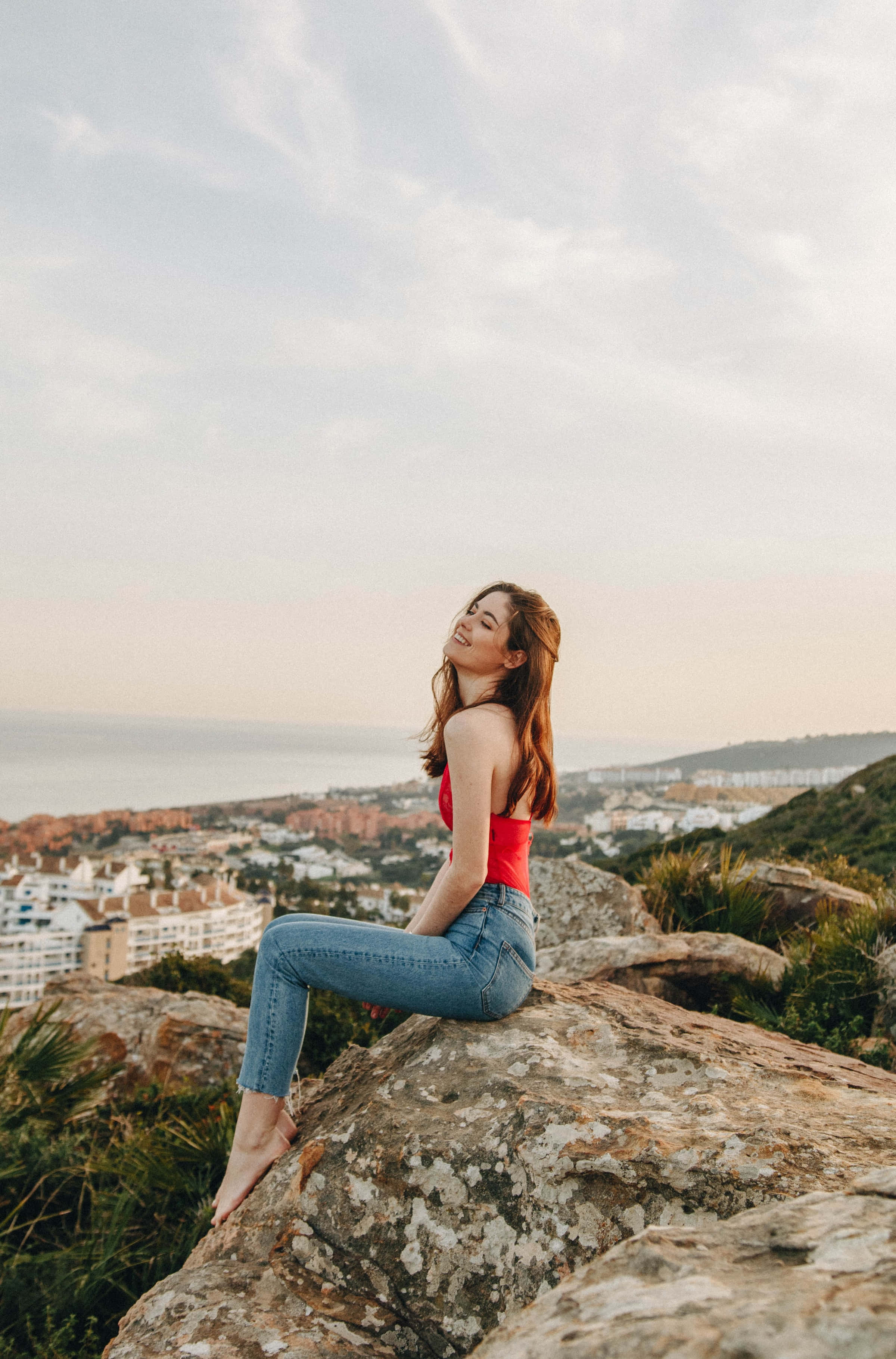 Woman Beautiful 桌布 壁紙 — 一位身穿紅色上衣和牛仔褲的女性赤腳坐在岩石懸崖上，俯瞰日落時分的沿海小鎮。