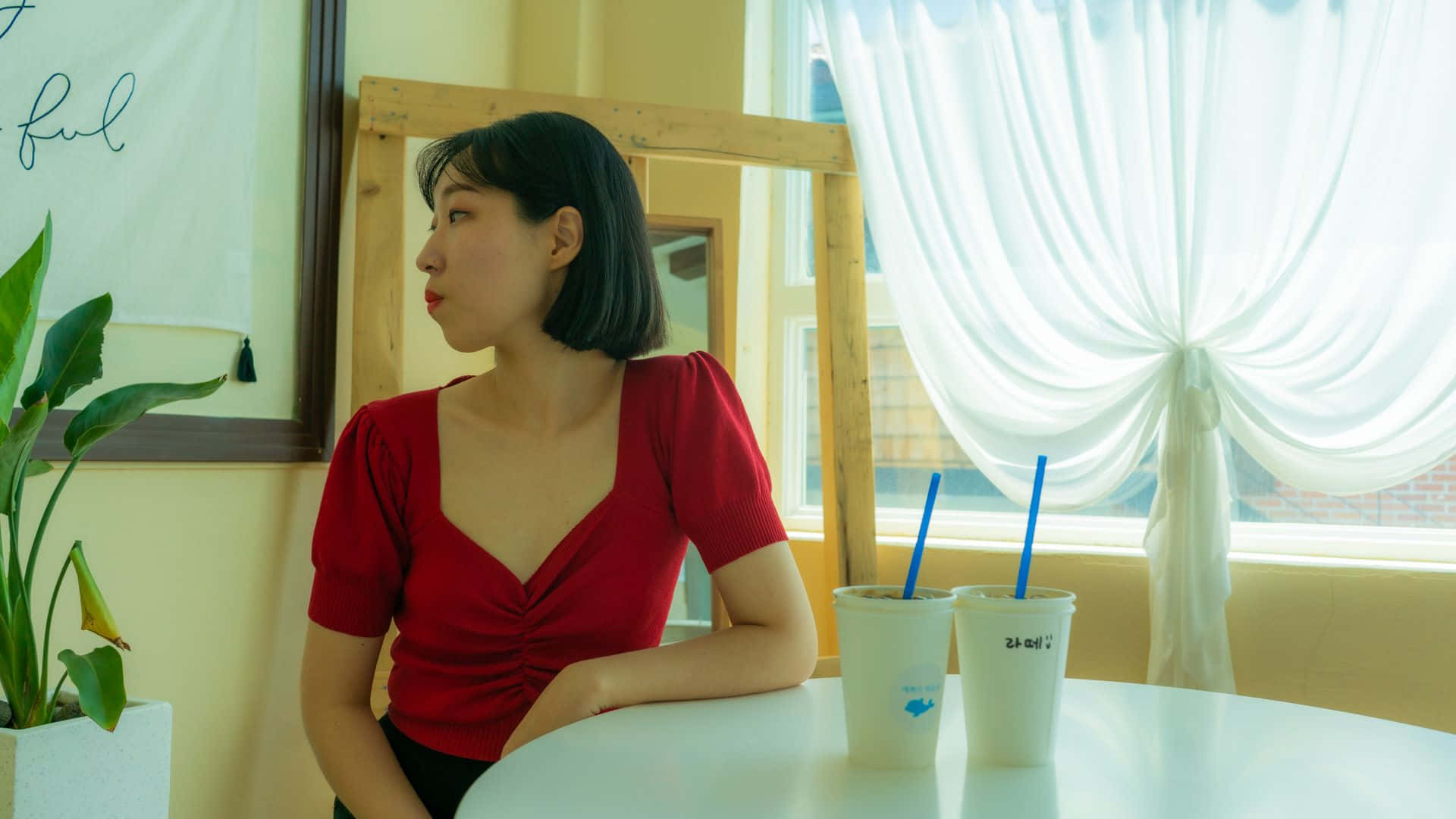 K-pop 桌布 壁紙 — 一位身穿紅色上衣的女性坐在窗邊的白色桌子旁，窗簾是透明的，旁邊有兩杯飲料。