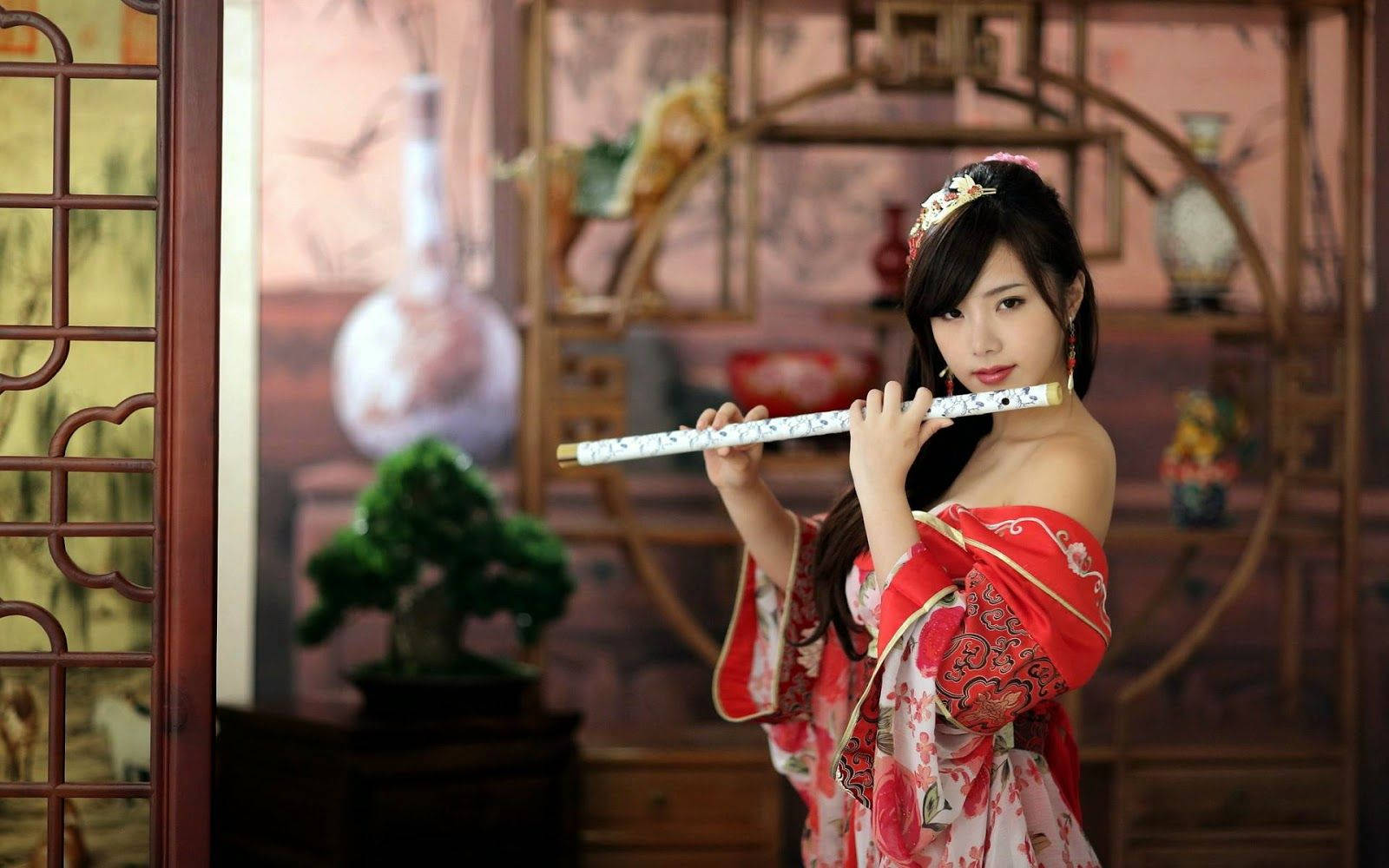 Sexy Model 桌布 壁紙 — 一位身穿紅色傳統長袍的女性在一個帶有裝飾架和盆景的木製室內吹奏長笛。