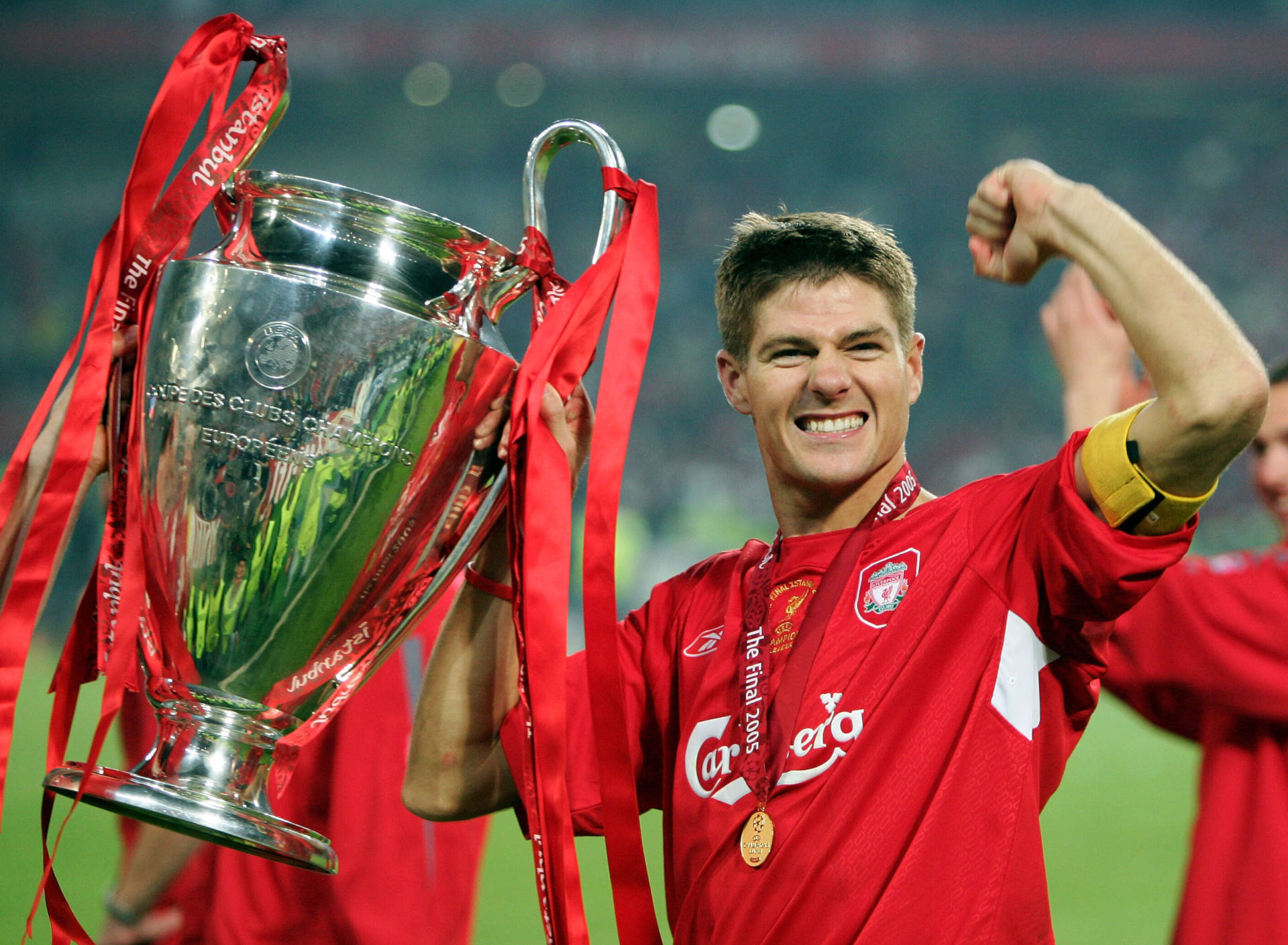 Steven Gerrard 桌布 壁紙 — 一位身穿紅色利物浦球衣的歡欣足球員，勝利地高舉閃閃發光的歐冠獎盃，獎盃上繫著紅色絲帶。