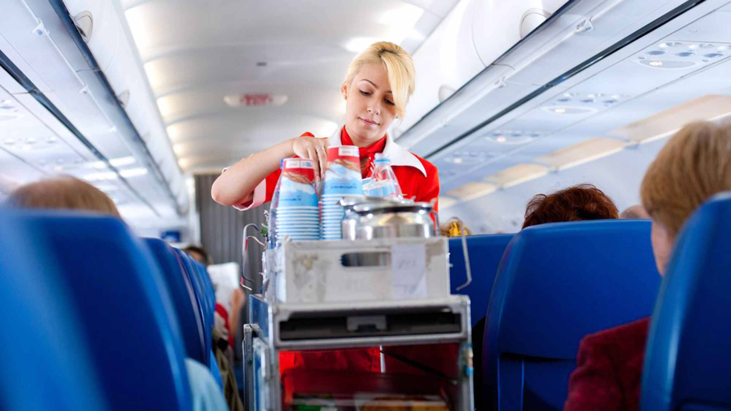 Airport 桌布 壁紙 — 一位身穿紅色制服的空服員正從手推車上提供飲料，沿著光線明亮的飛機客艙通道，座位為藍色。