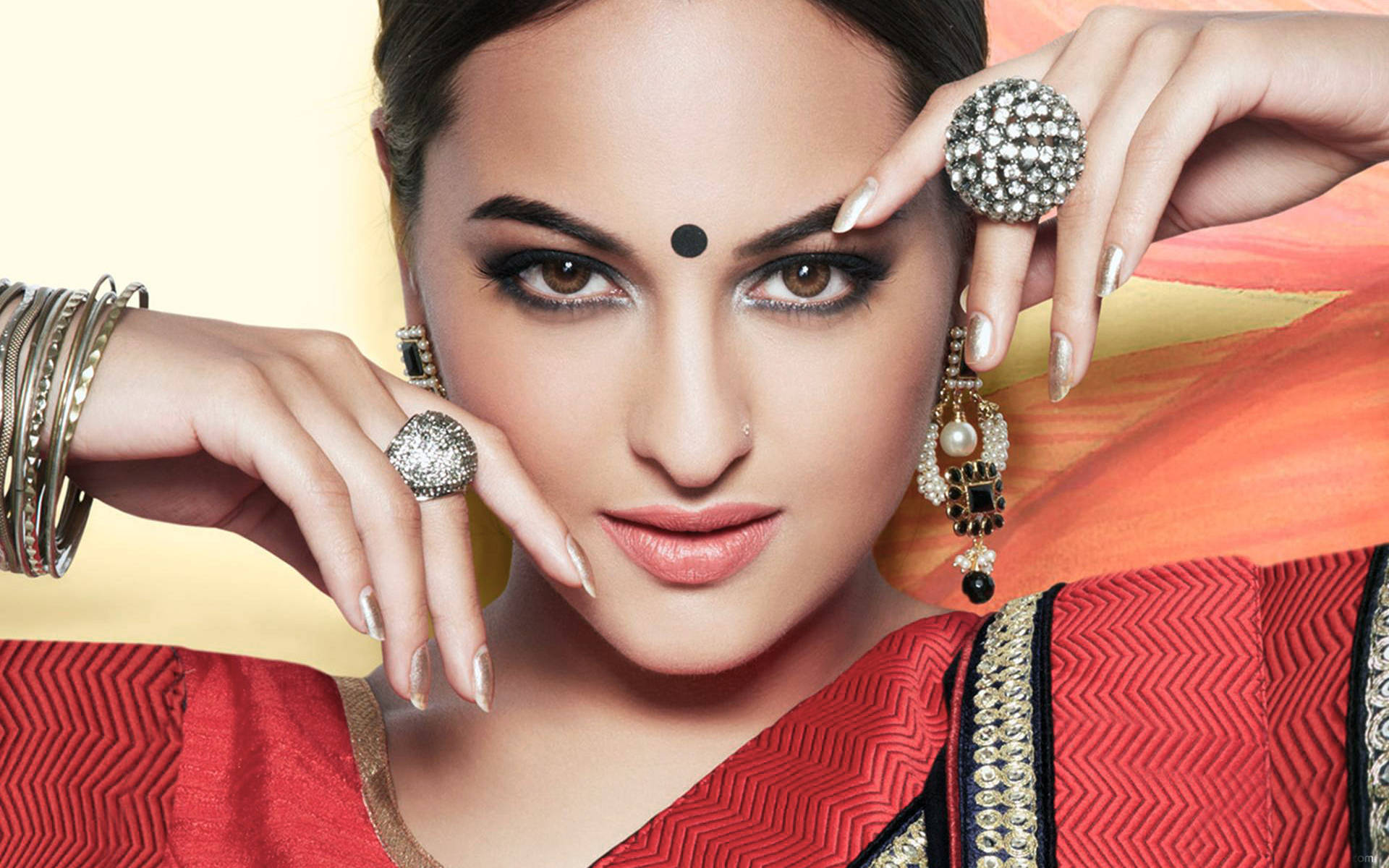 Sonakshi Sinha 桌布 壁紙 — 一位身穿紅色圖案紗麗、飾以精緻珠寶的女性，在溫暖漸變背景前自信地擺姿勢。