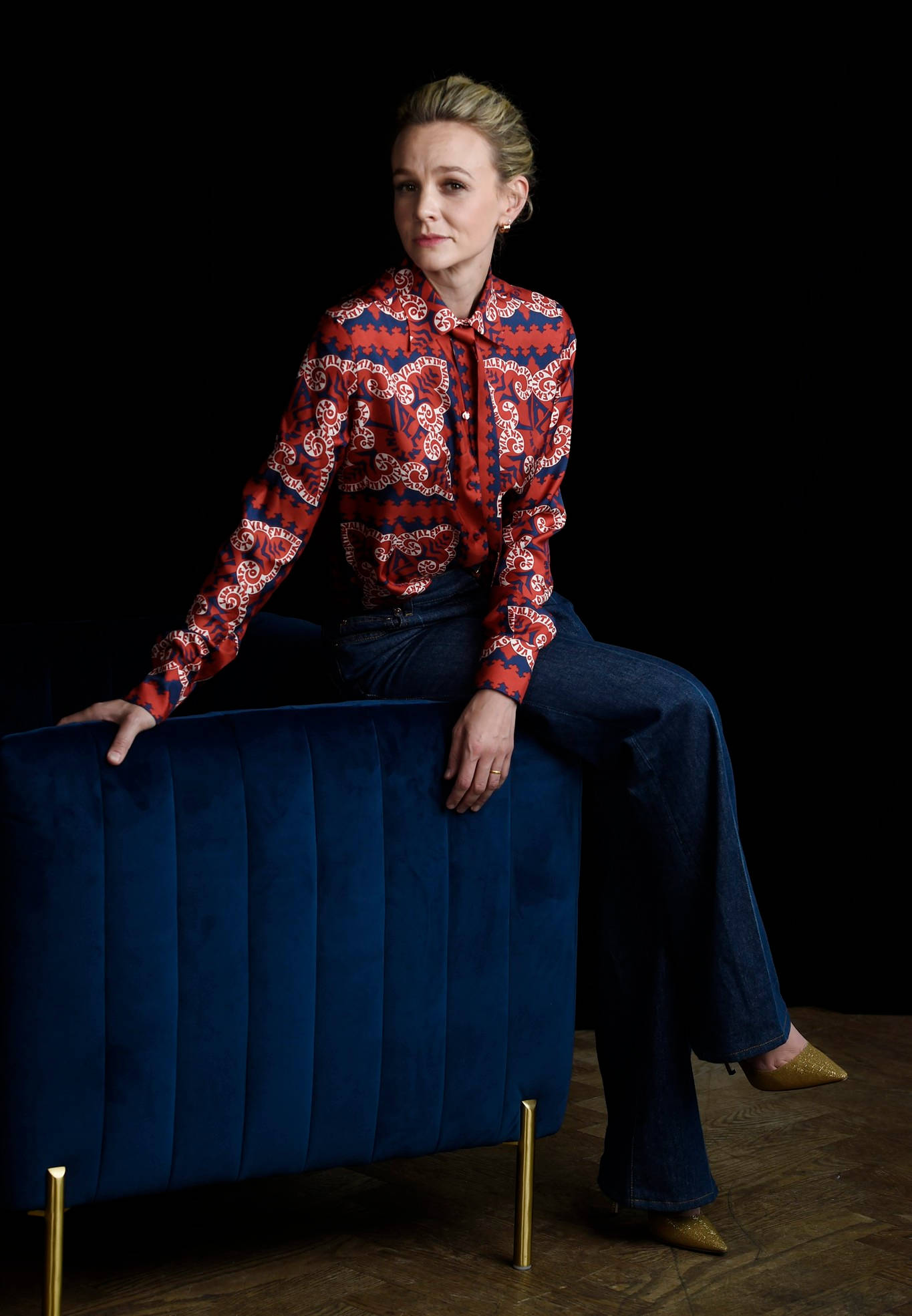 Carey Mulligan 桌布 壁紙 — 一位身穿紅色圖案襯衫和藍色牛仔褲的女性，在純黑背景下，端坐在深藍色天鵝絨腳凳上。