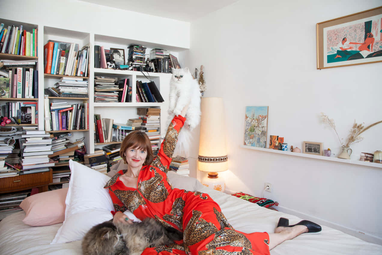 Bedroom 桌布 壁紙 — 一位身穿紅色圖案長袍的女性躺在床上，高舉一隻白貓，一隻灰貓在她旁邊休息，周圍是書架和溫暖的燈光。