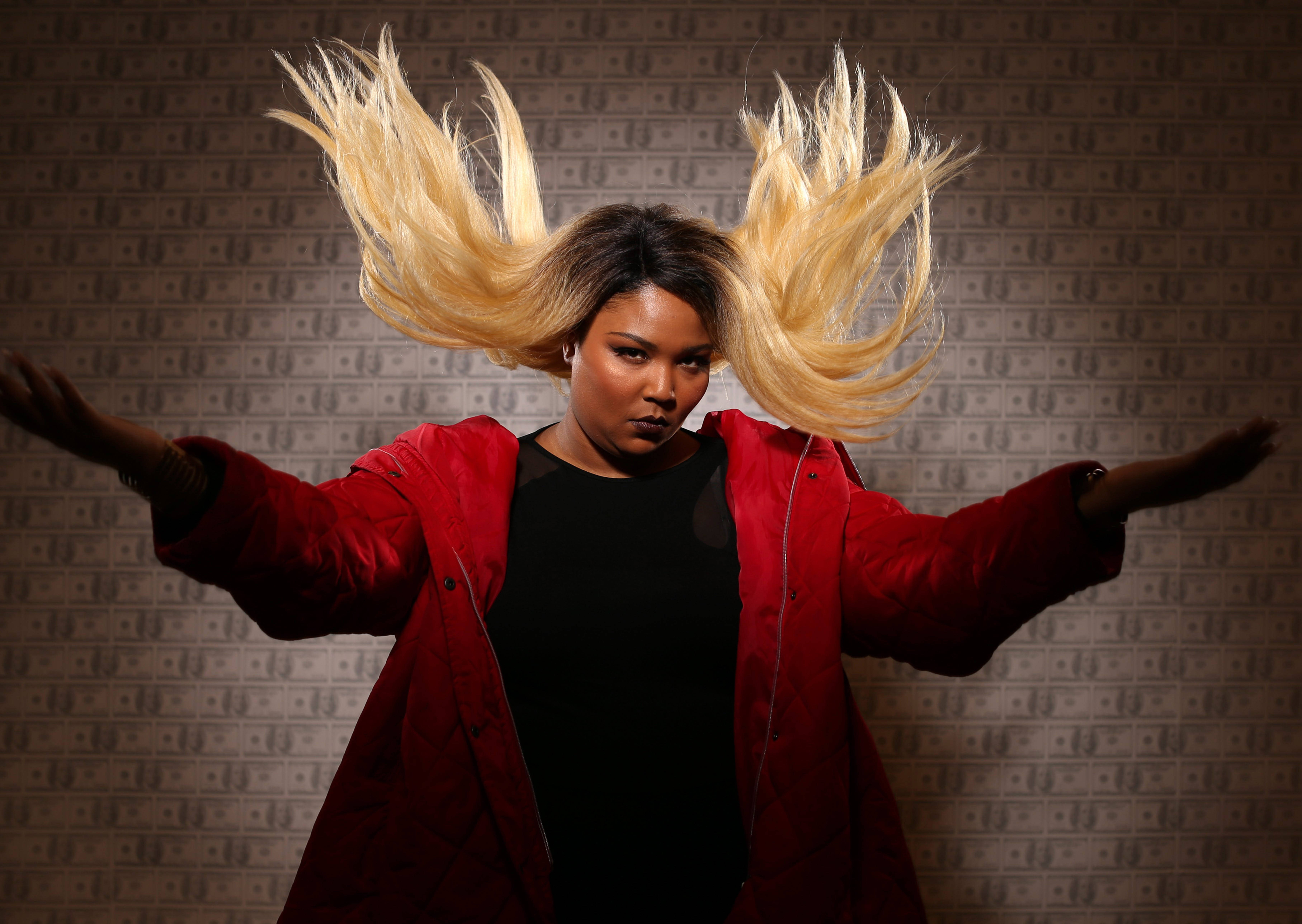 Lizzo 桌布 壁紙 — 一位身穿紅色外套、金髮飄揚的女性，在美元鈔票壁紙背景前戲劇性地飛舞。
